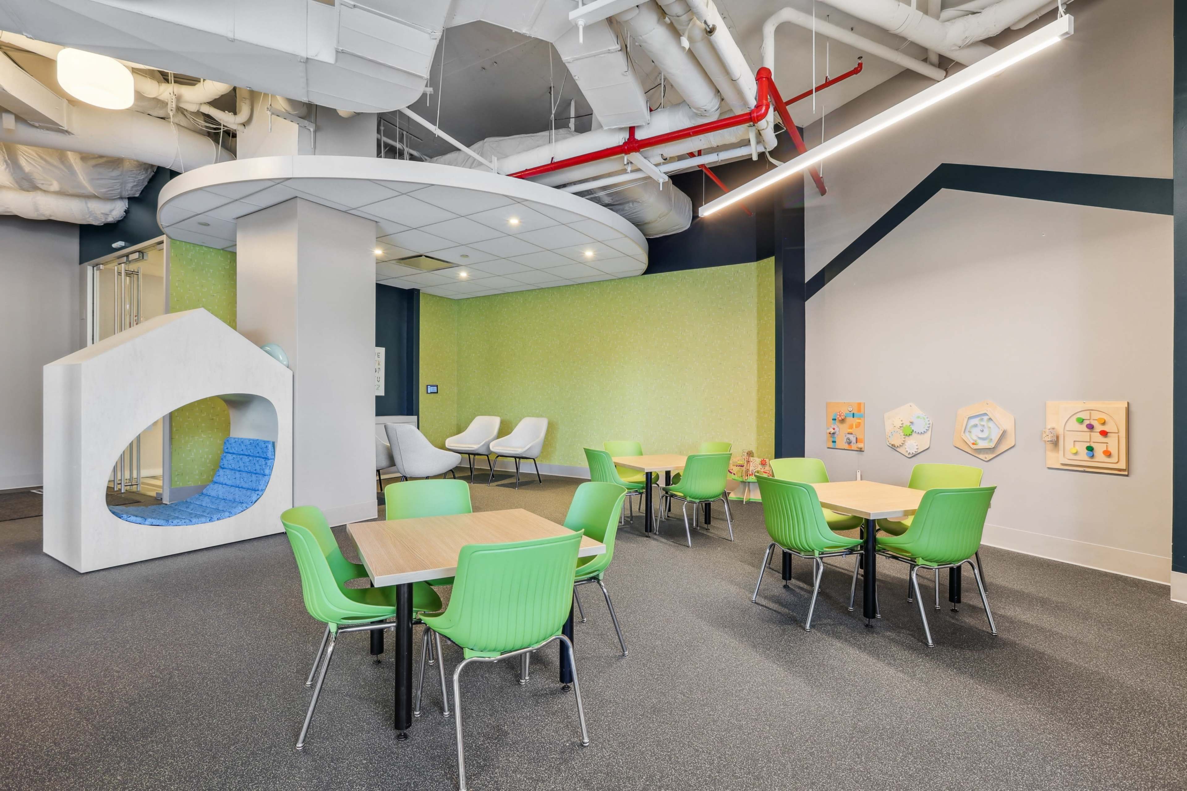 The image depicts a modern indoor play area featuring several tables with green chairs, a white play structure, and a colorful wall with art displays.