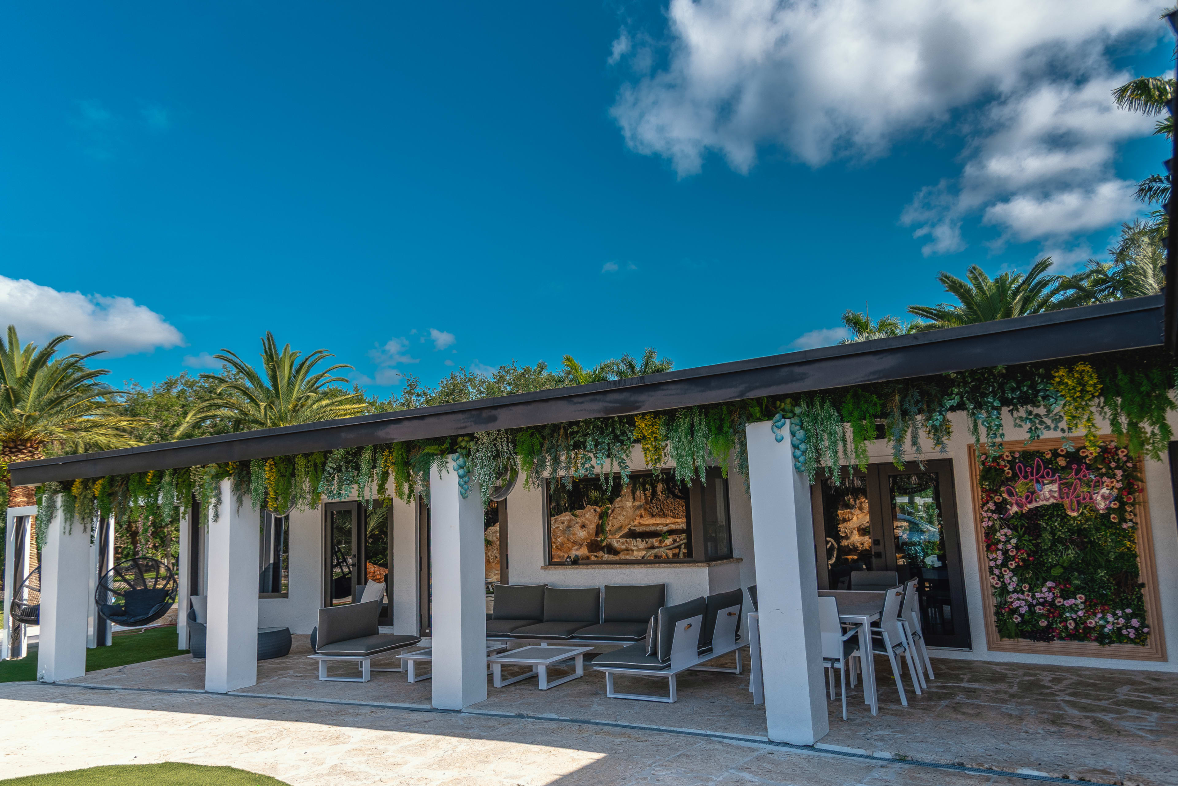 A modern outdoor lounge area features white furniture and a wall adorned with greenery and floral decorations, set against a backdrop of palm trees and a bright blue sky.