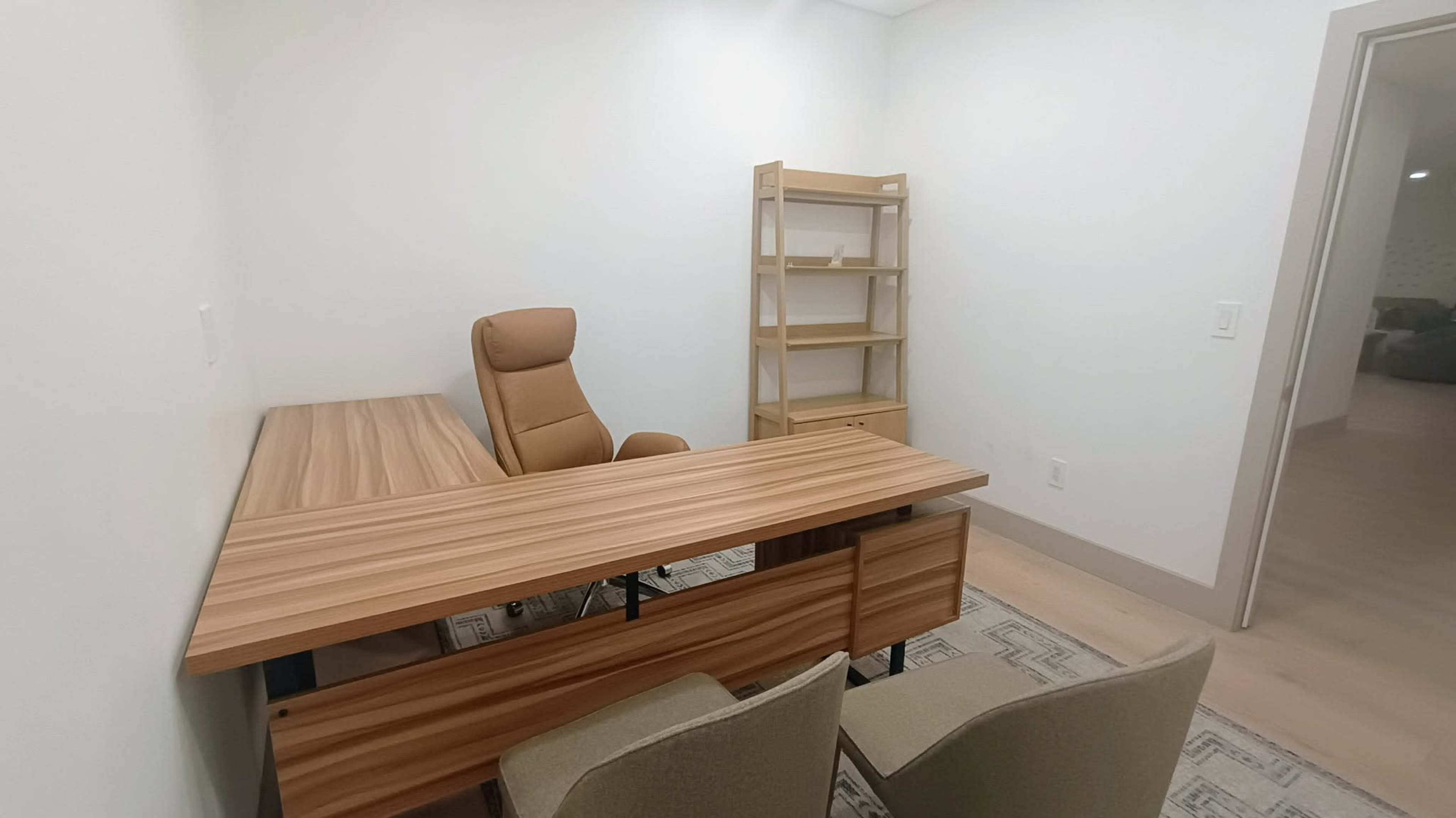 The image shows a small, empty office space featuring a wooden desk with a padded chair and a shelf against the wall.