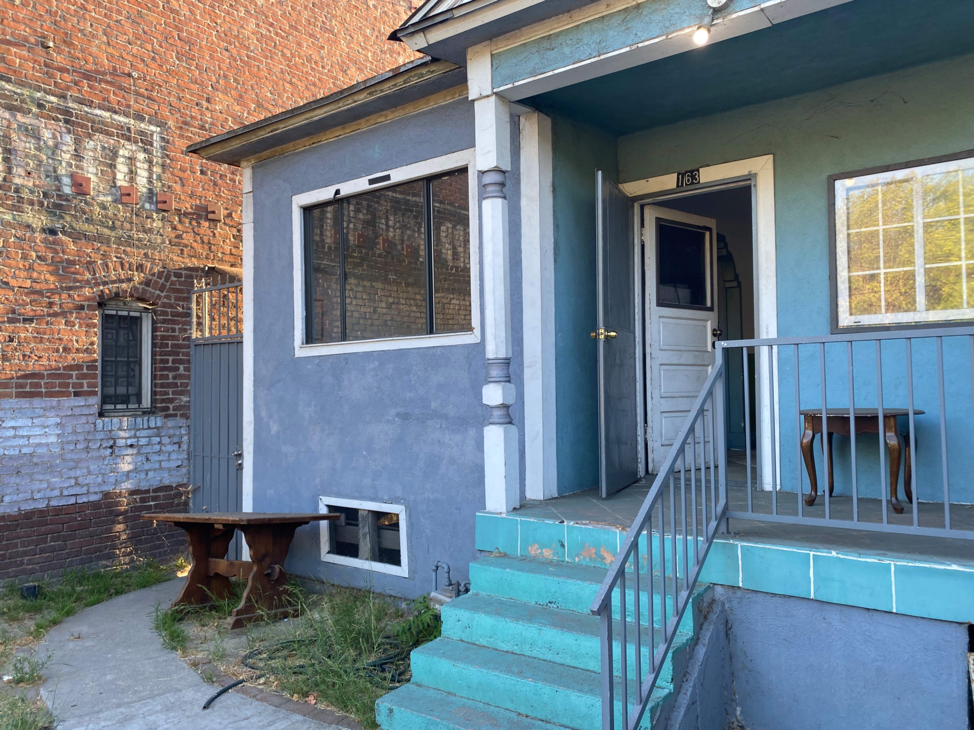 A blue house with a porch, a front door, and a small table visible next to the entrance, situated next to a brick wall.