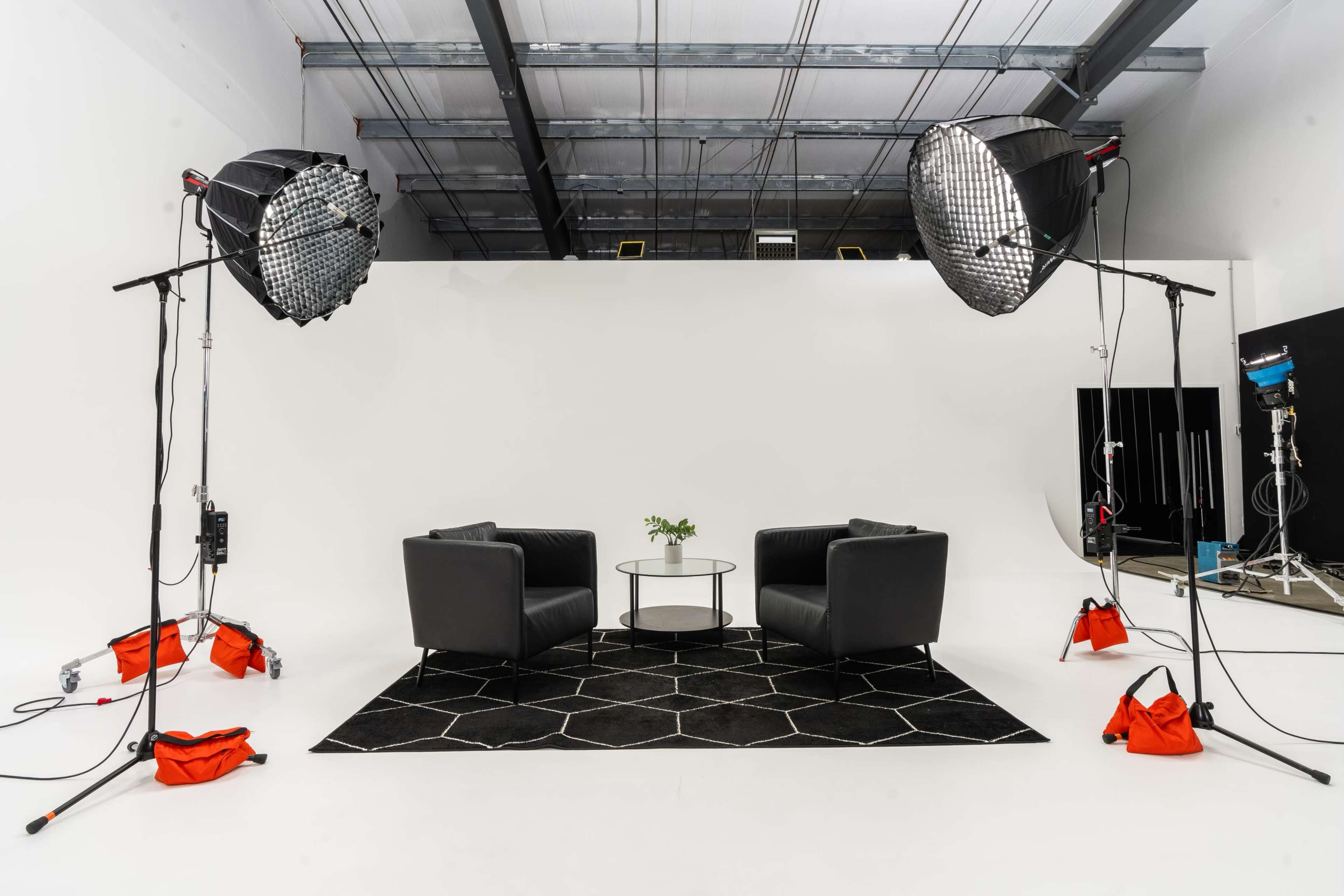 A minimalist photography studio setup with two black chairs facing each other, a glass table in the center, and large softbox lights positioned on either side.