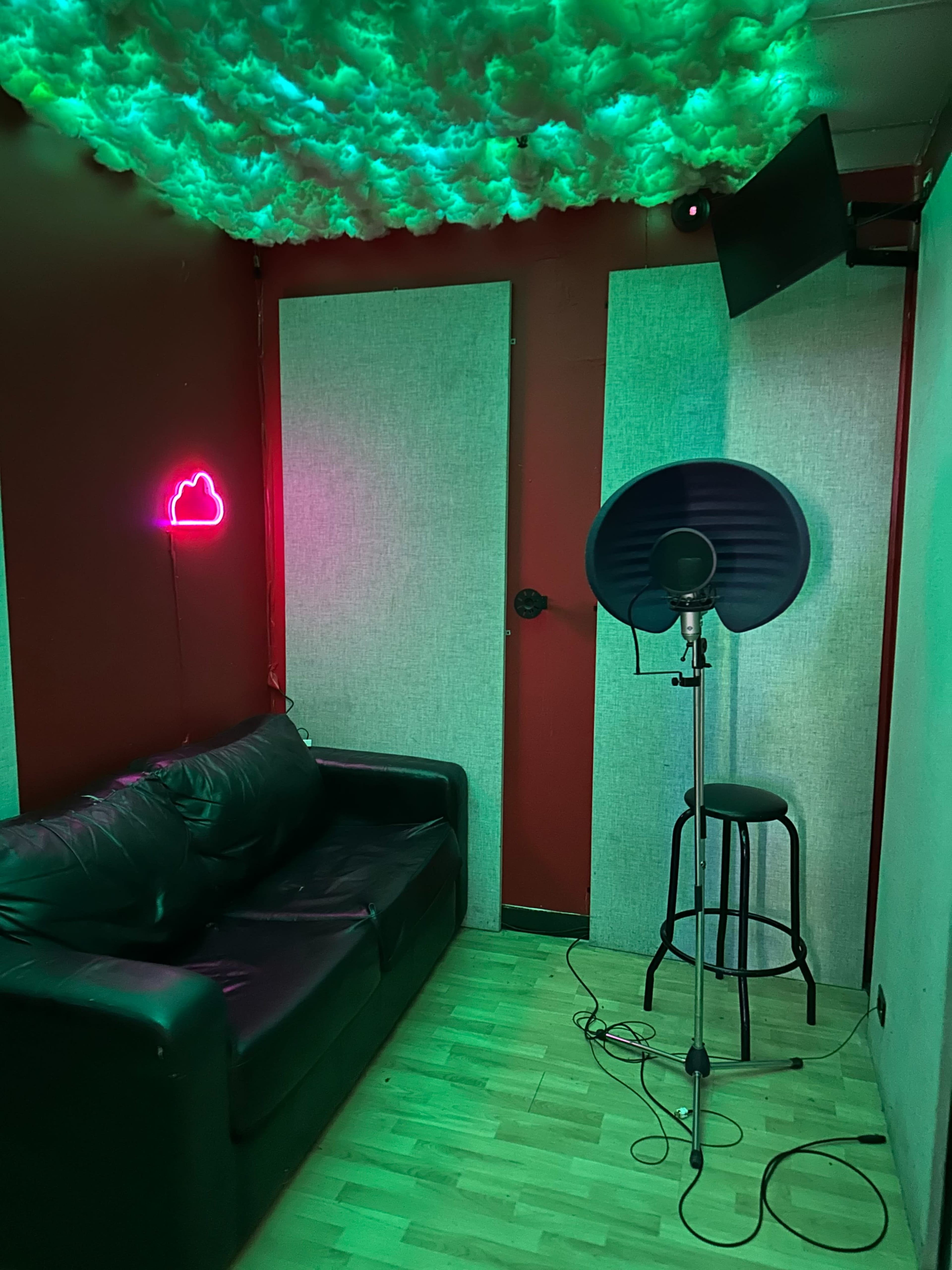 The image shows a soundproof recording booth featuring a black couch, a microphone on a stand, a stool, and green lighting with a neon cloud sign.