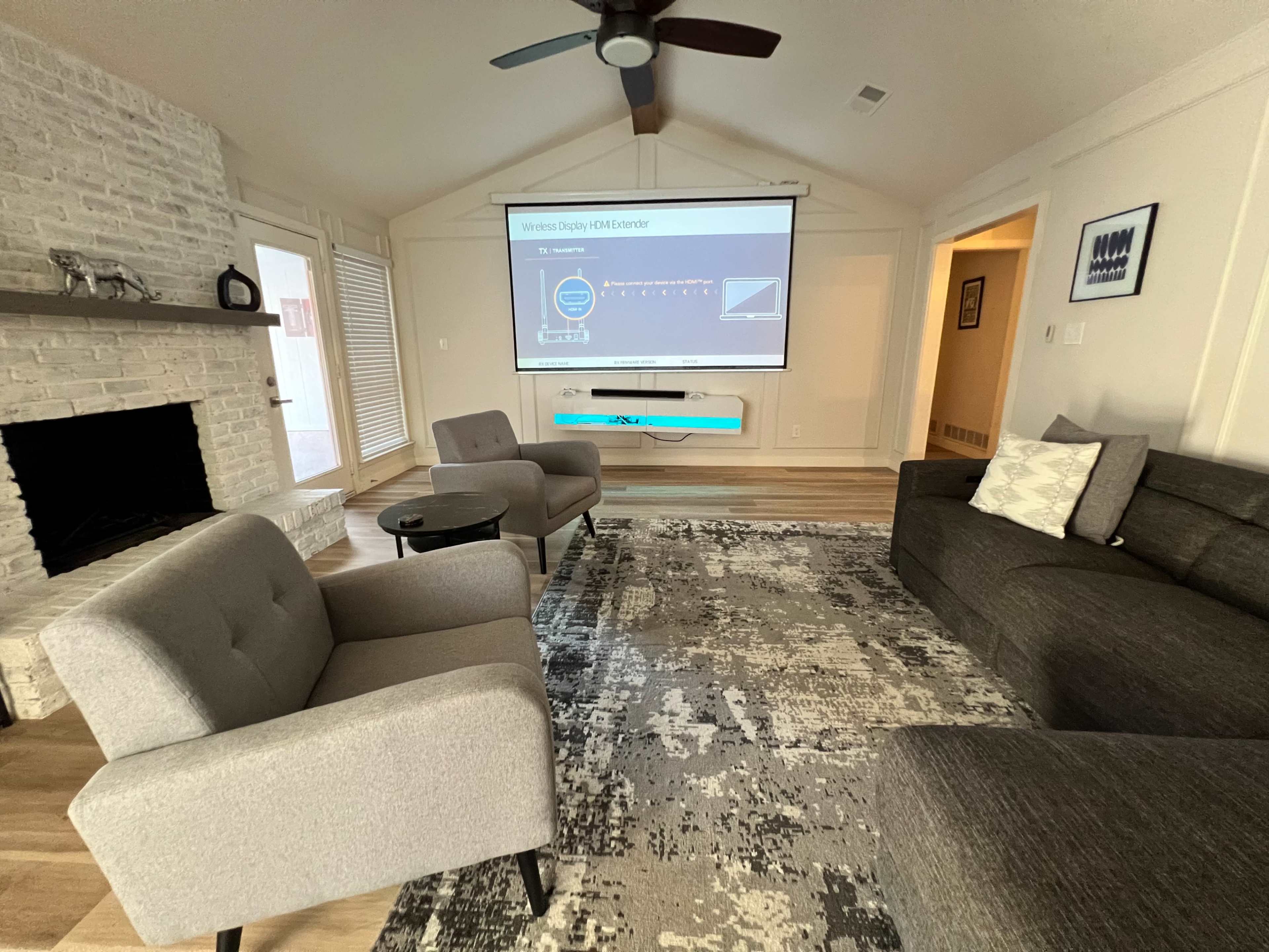 A modern living room with a large screen displaying a presentation, two gray upholstered armchairs, a gray sofa, and a patterned area rug on wooden flooring.