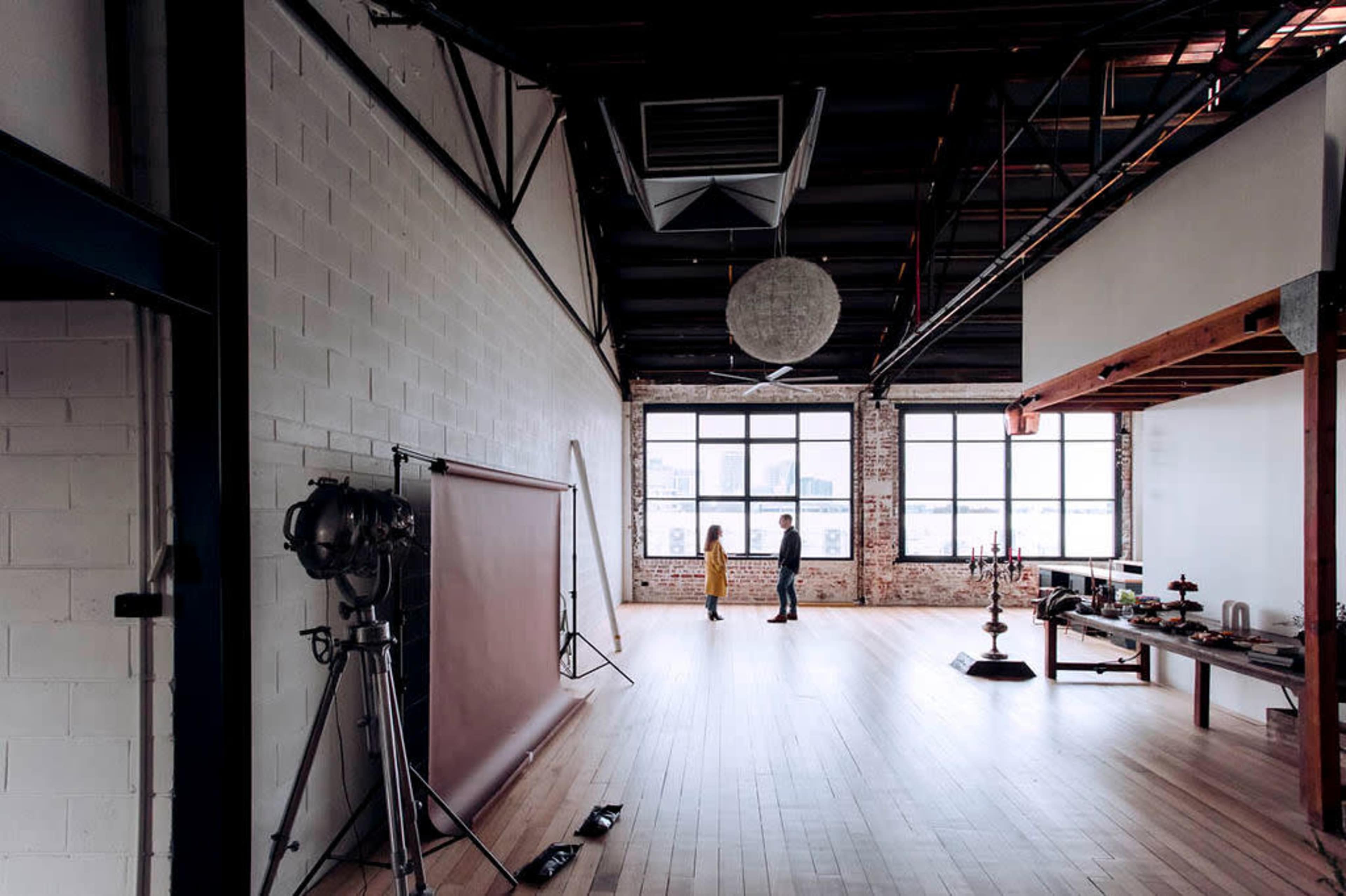Two people stand in a spacious, industrial-style room with large windows and wooden flooring, surrounded by photography equipment and furnishings.
