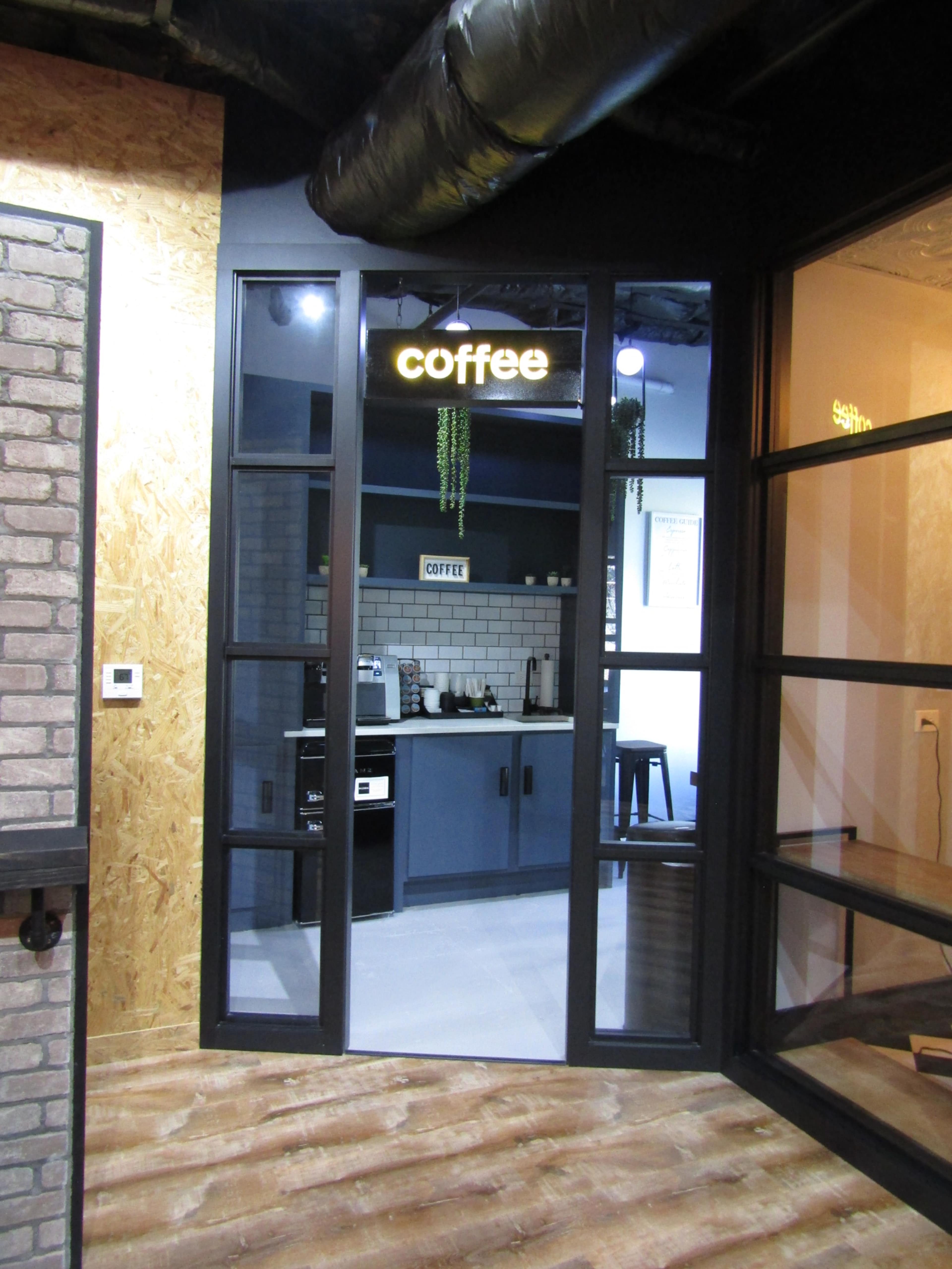The image shows a modern office space with a glass door labeled "coffee" leading to a kitchen area featuring a coffee station.