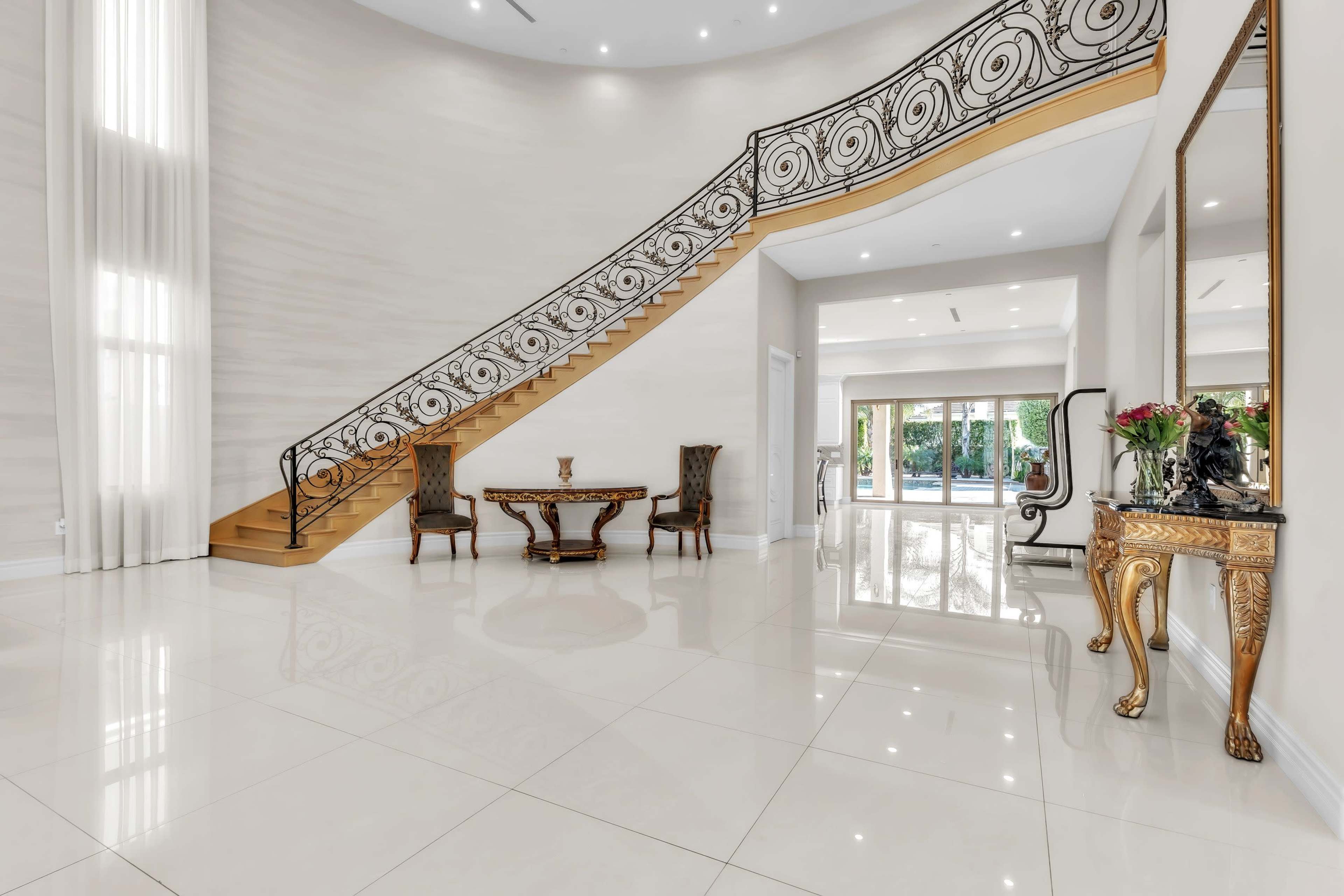 The image shows a spacious entryway featuring a curved staircase with ornate black railing, a decorative table, and large windows that provide natural light.