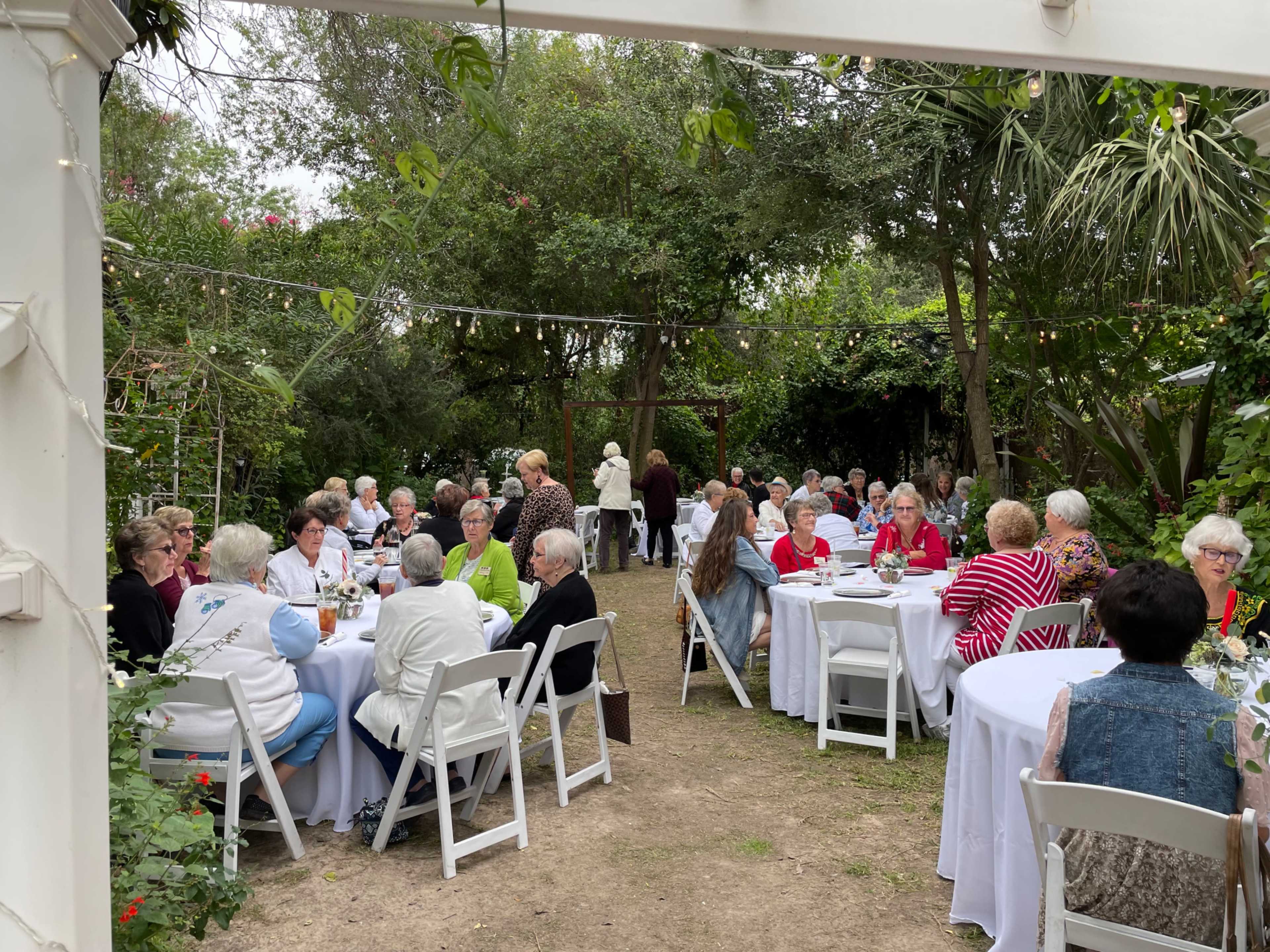 A group of people is seated at round tables in a garden setting, enjoying a meal amidst greenery and string lights.