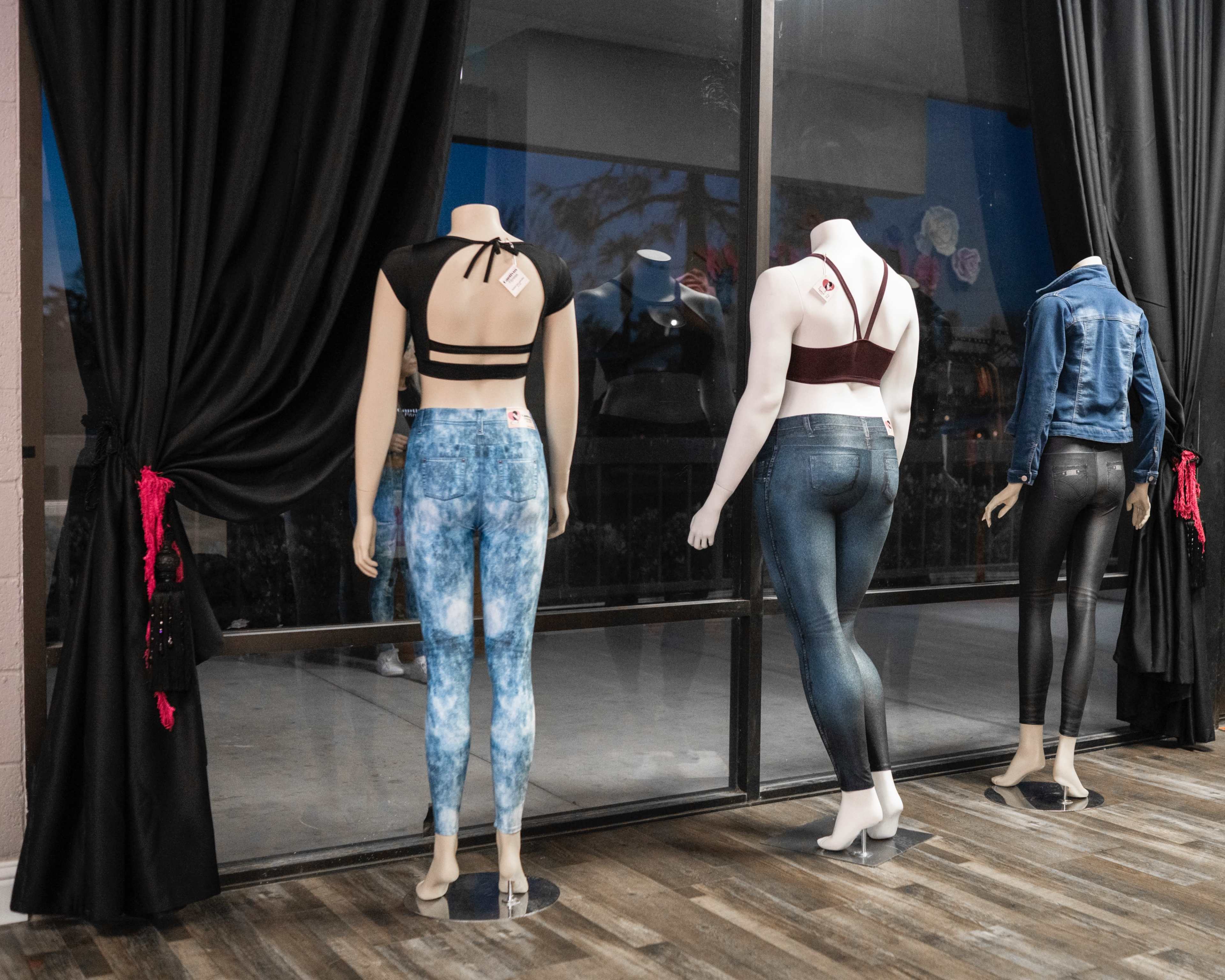 Three mannequins display various styles of women's clothing, including tops and jeans, in front of large windows with dark curtains.