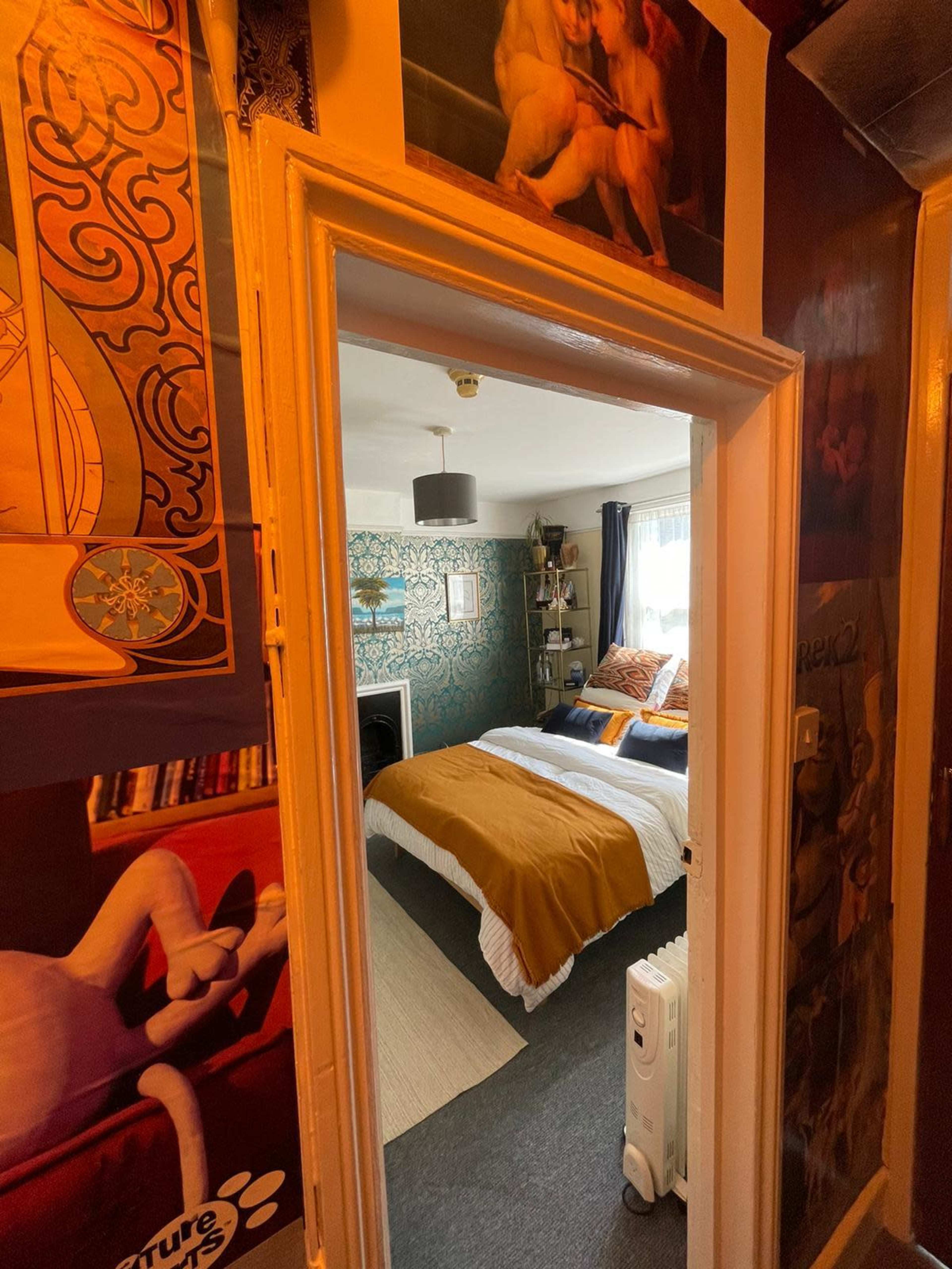 A doorway leads into a bedroom with a bed covered in a yellow and white bedspread, surrounded by decorated walls and a window.