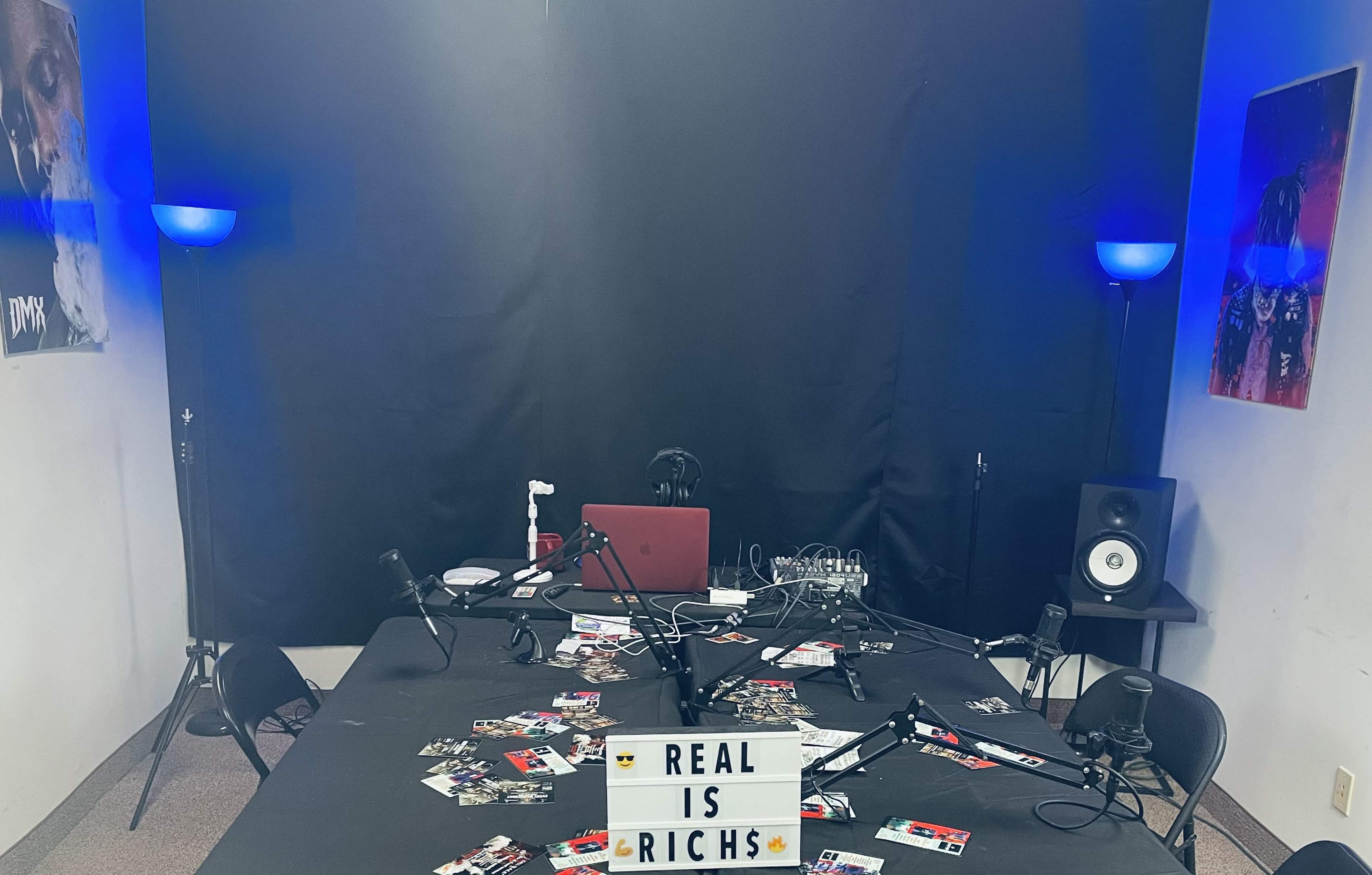 A recording studio with a black backdrop, multiple microphones, an array of scattered business cards, and a lit sign that reads "REAL IS RICHS."