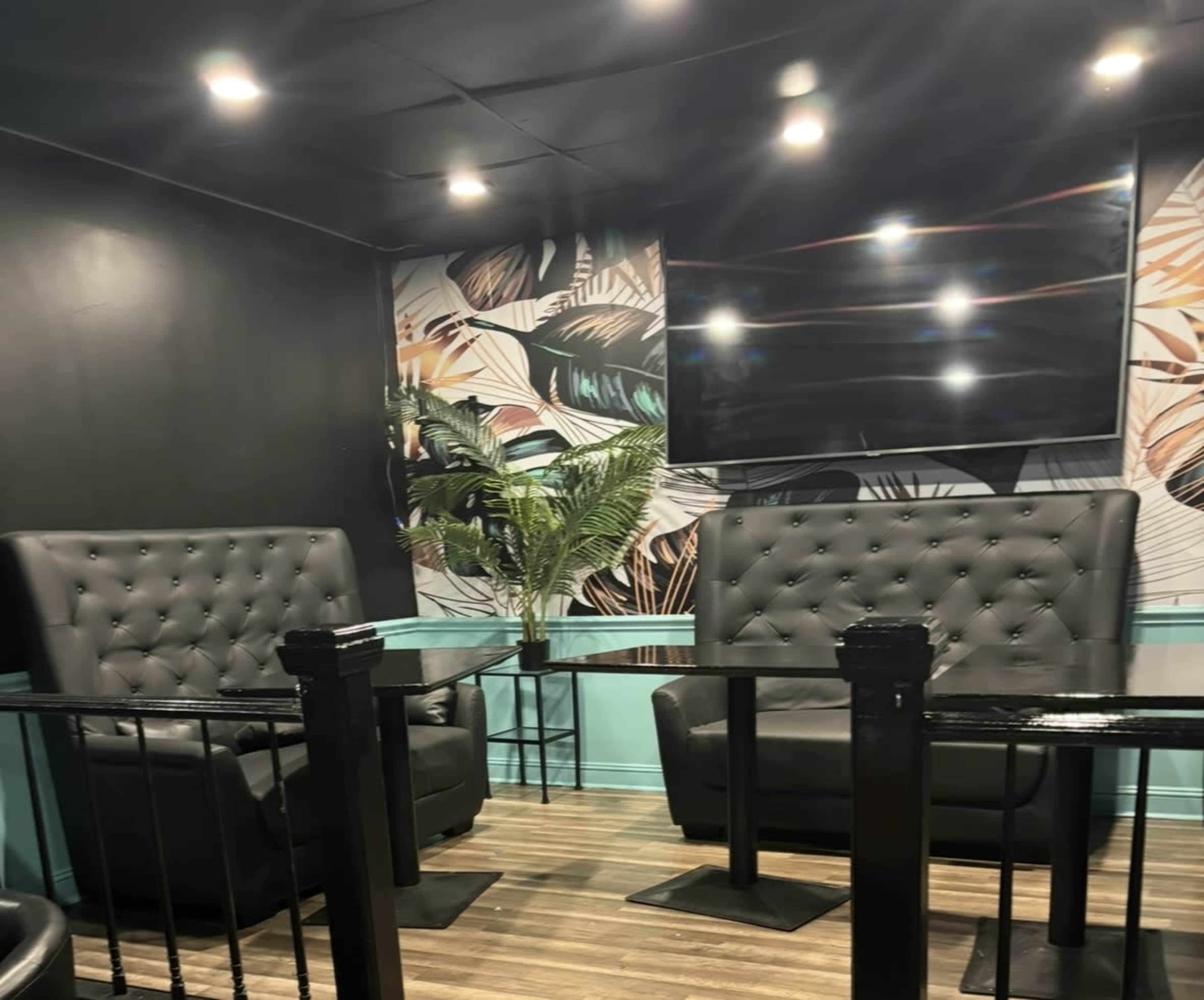 The image shows a cozy seating area with two black upholstered chairs, a small glass table, and a tropical-themed wallpaper.