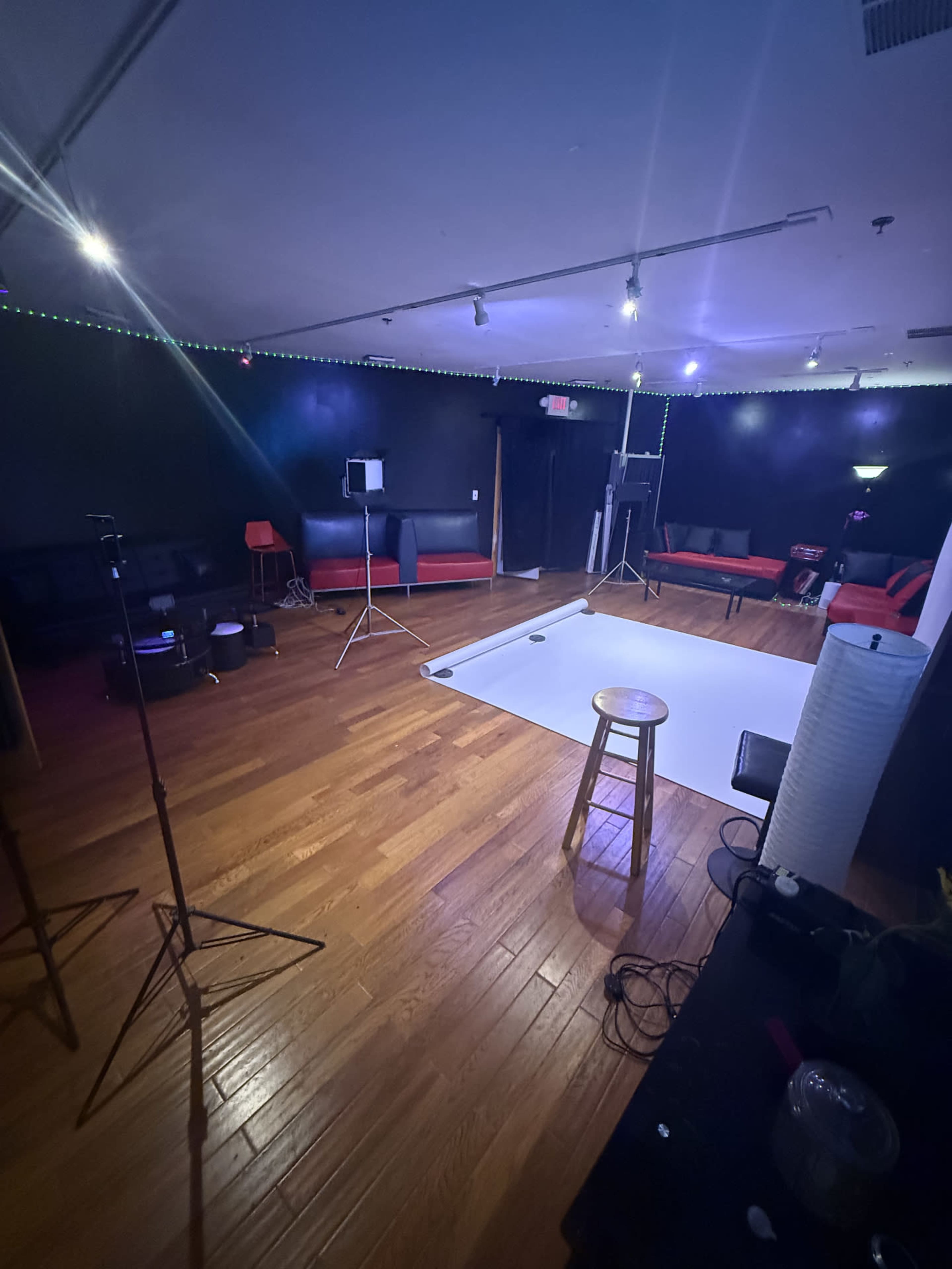 The image shows an empty photography studio with wooden floors, a white backdrop set up, and various lighting equipment positioned around the room.