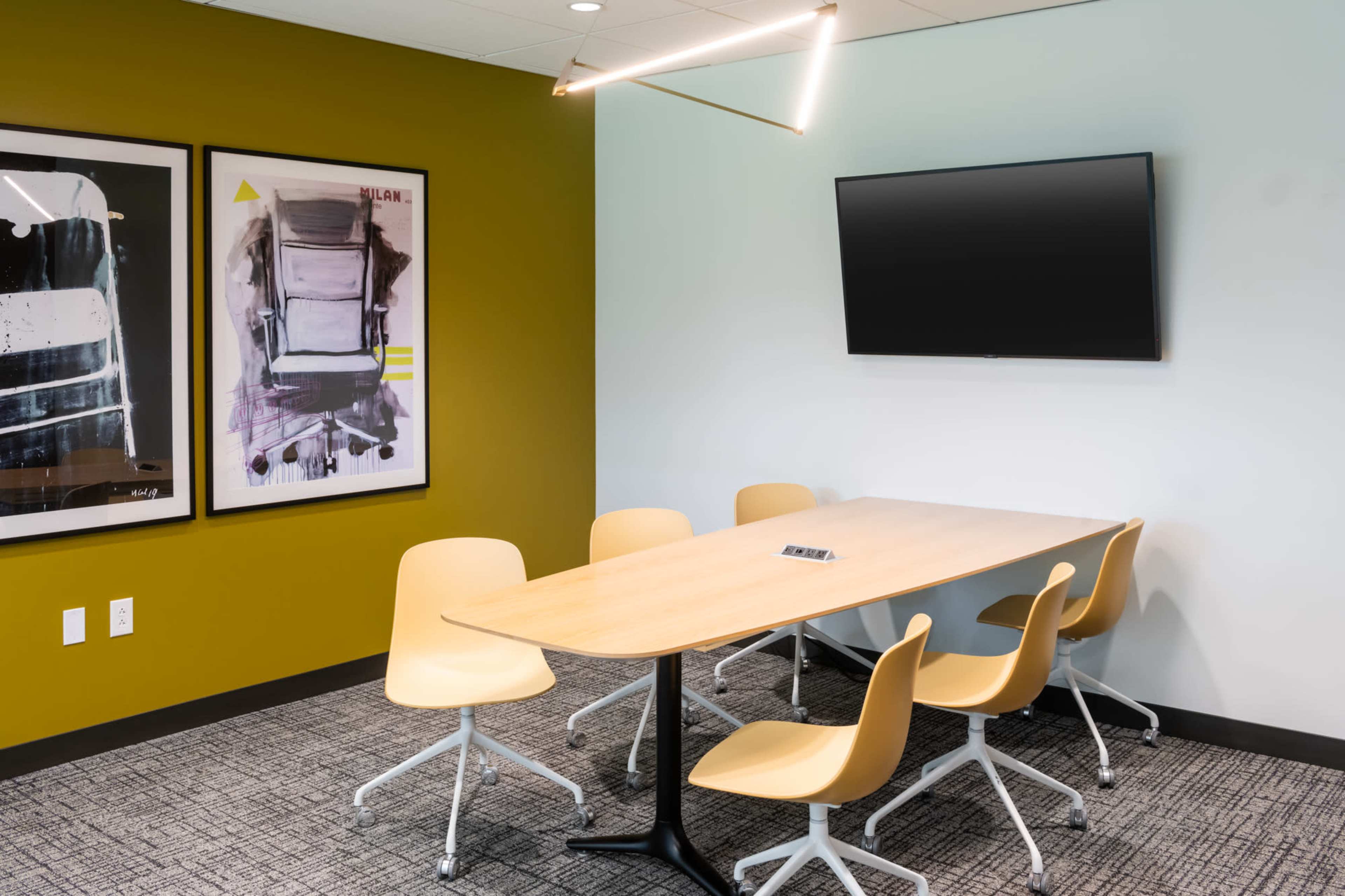 The image shows a modern conference room with a wooden table, six yellow chairs, a television on the wall, and two framed artworks.