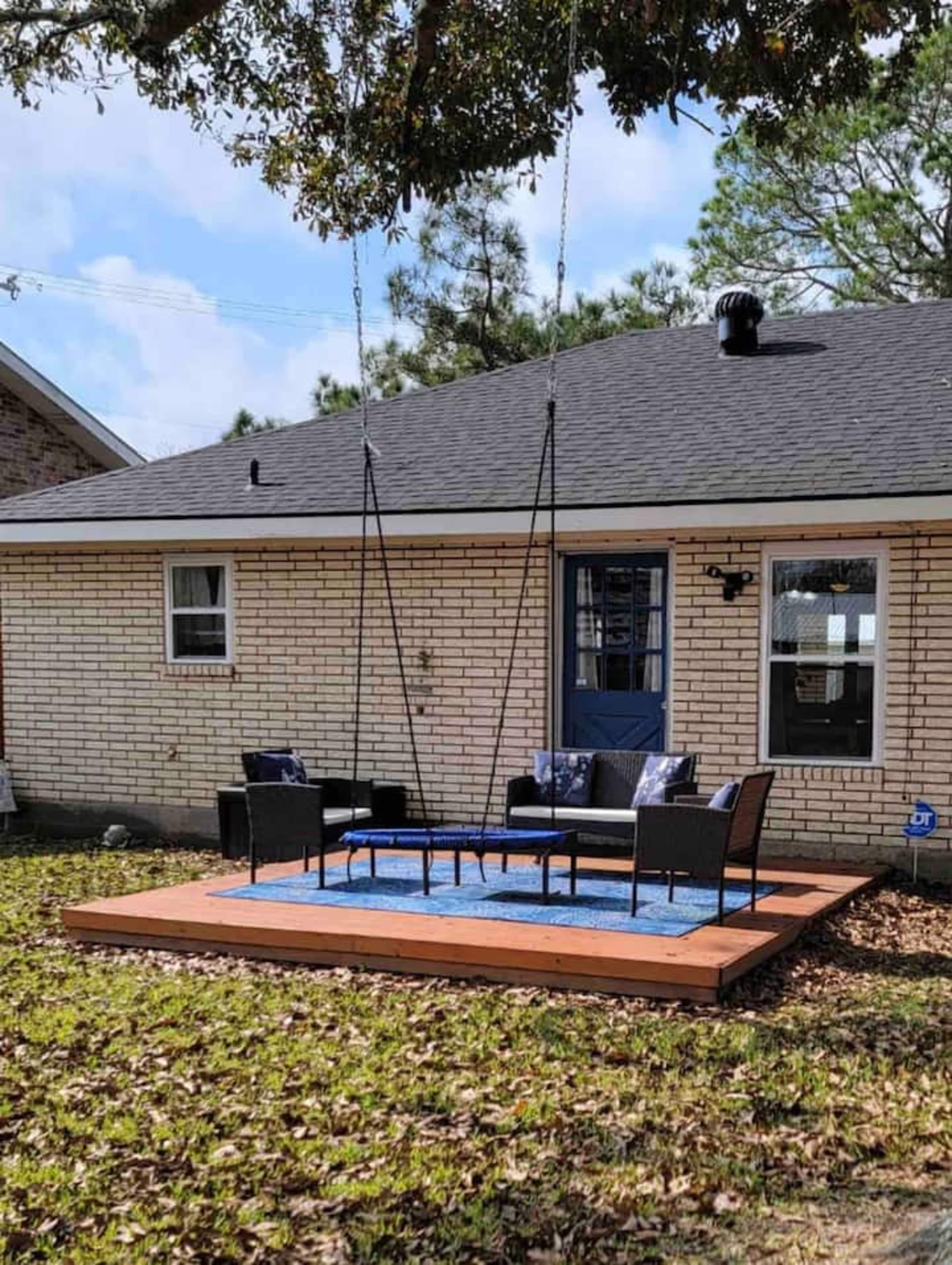 A wooden platform with a blue rug hosts a seating area of black wicker furniture and a table, located in a yard beside a brick house.