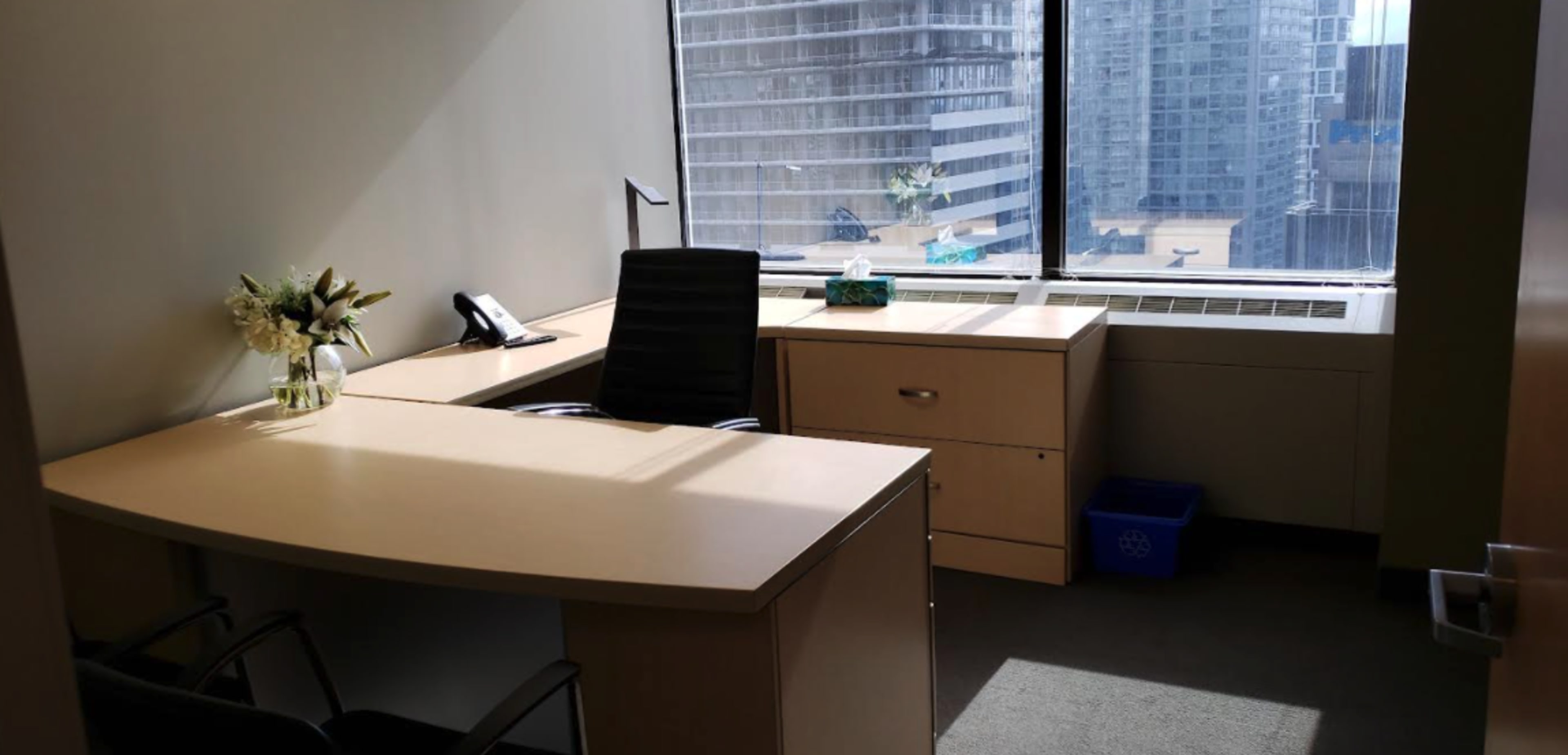 The image shows a modern office space with a desk, chair, and window displaying a view of tall buildings outside.
