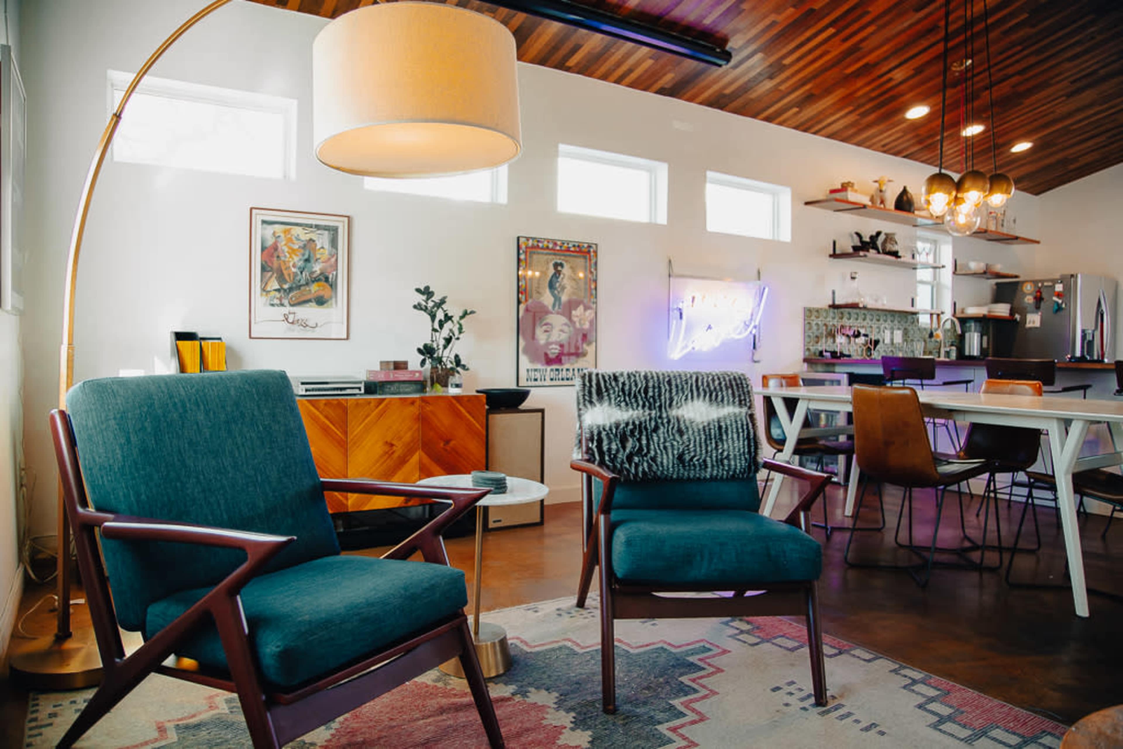 The image shows a modern living space featuring two teal chairs, a floor lamp, and a dining area with a bar in the background.