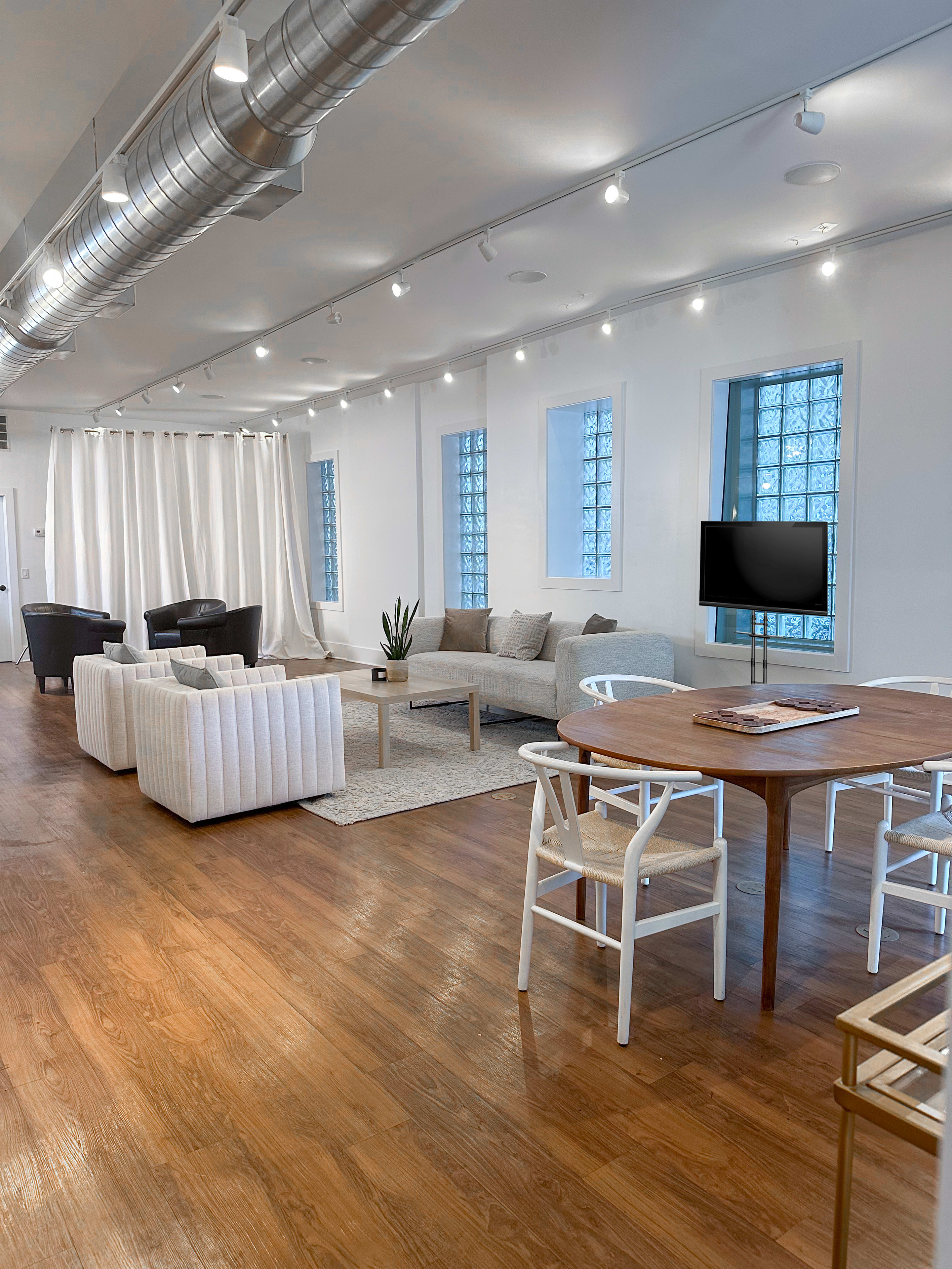 The image shows a modern open-concept living space featuring a seating area with white sofas, a round wooden dining table, and large windows with sheer curtains.