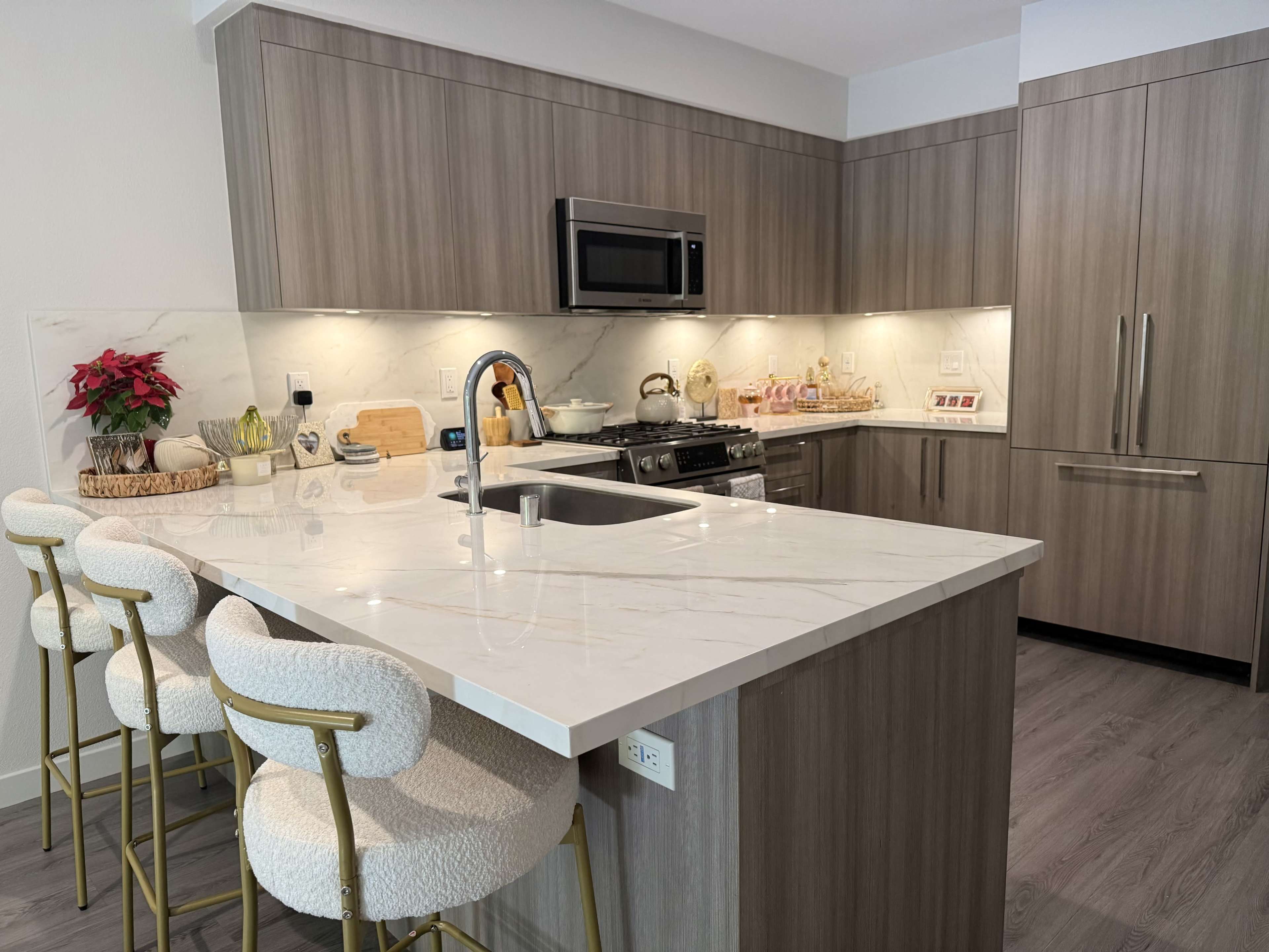 The image shows a modern kitchen featuring a marble countertop, stainless steel appliances, and wooden cabinetry, with three bar stools positioned at the island.