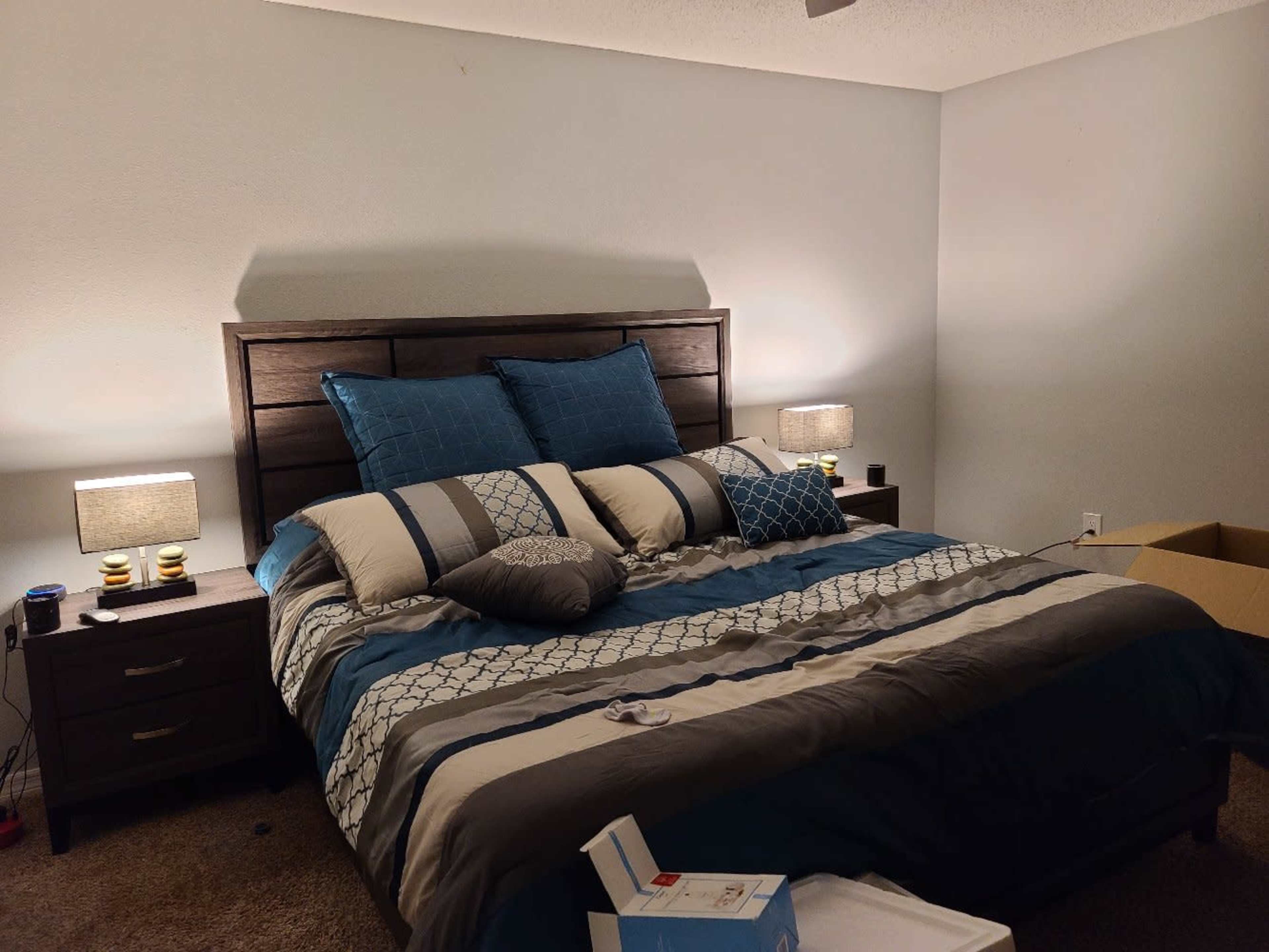 A neatly made bed with blue and gray bedding is centered in a softly lit bedroom, flanked by two bedside tables with lamps.