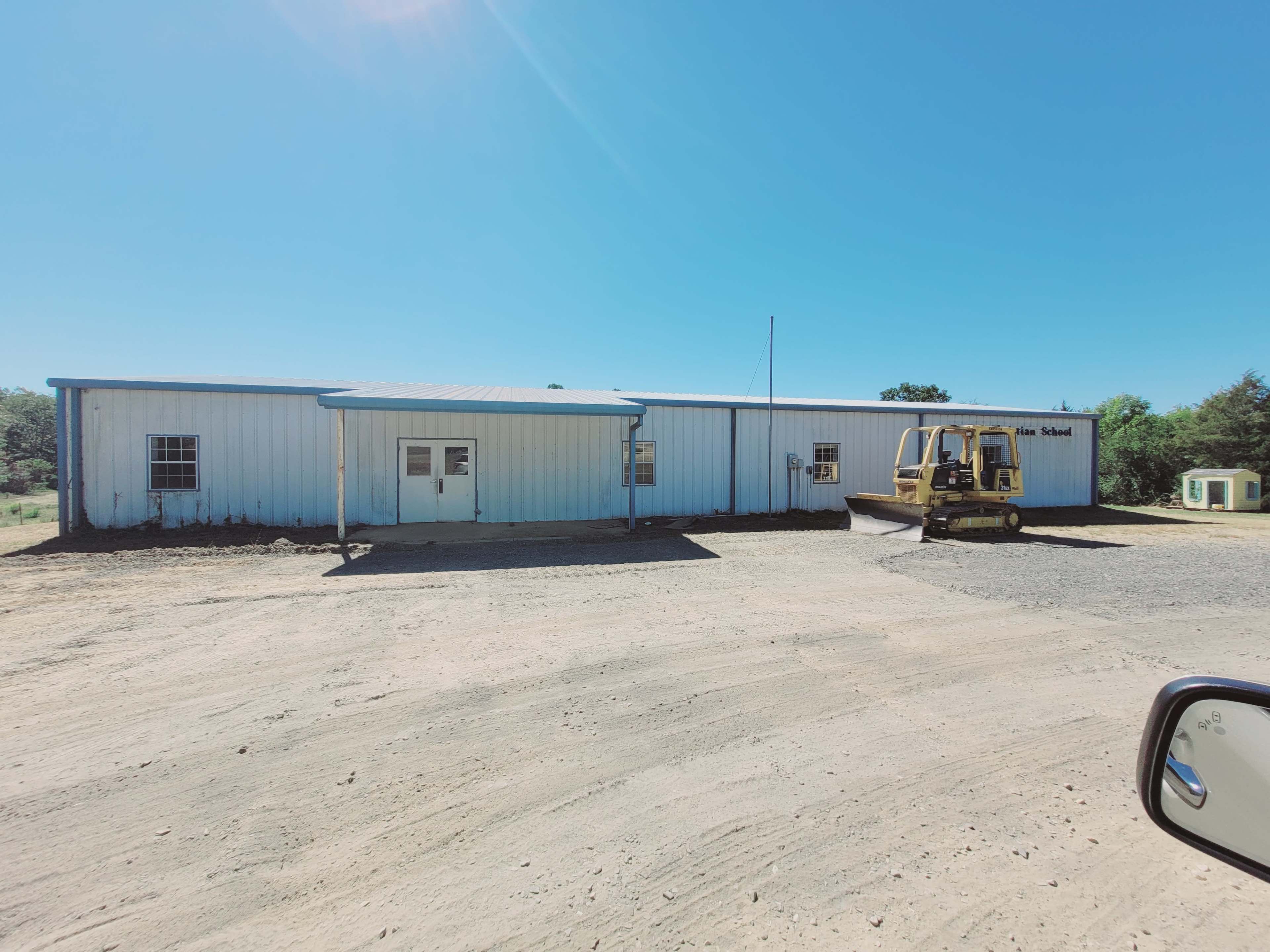 A large, single-story metal building with a blue roof stands on a gravel lot, accompanied by heavy machinery in front.