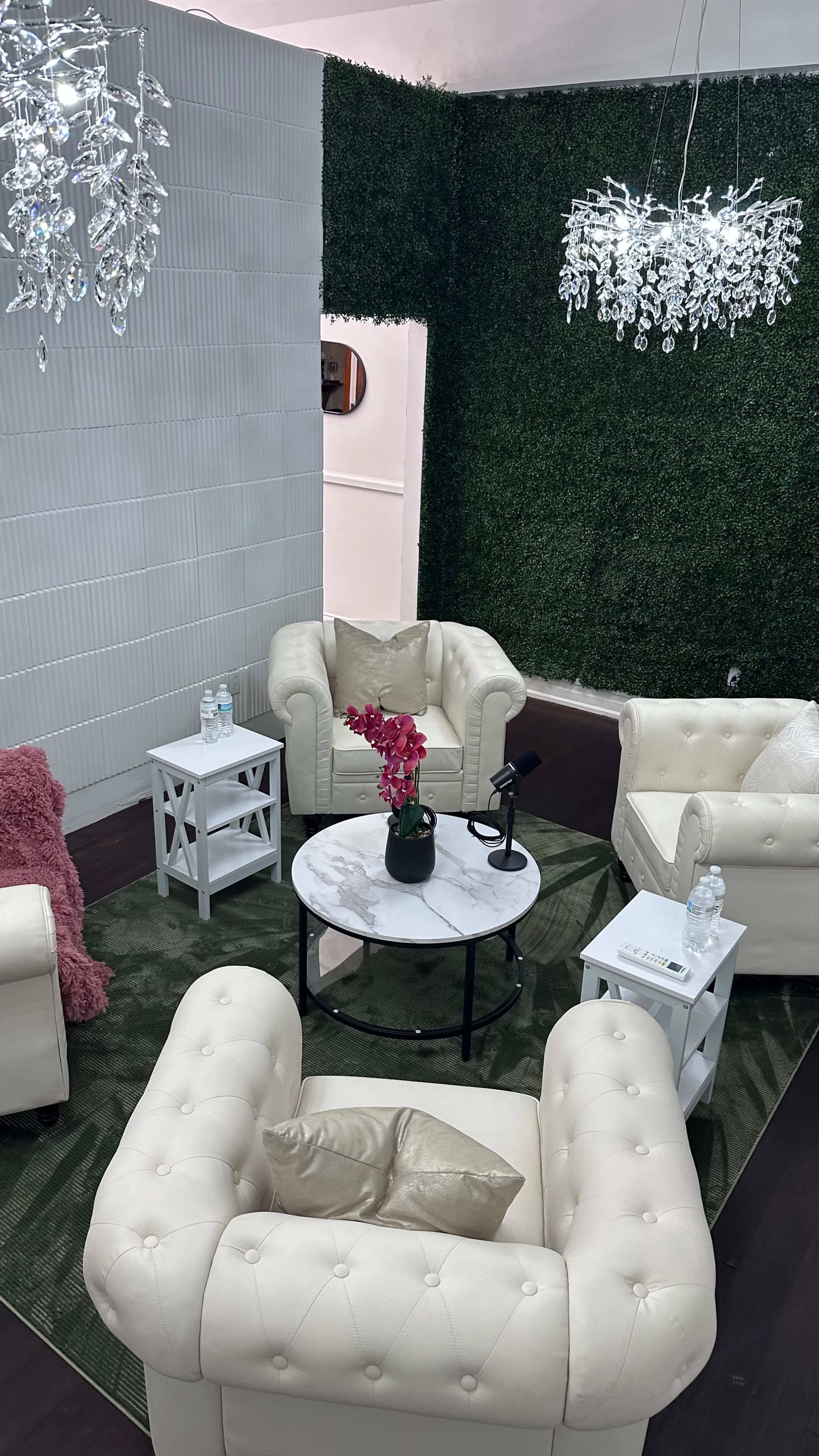 The image shows a cozy lounge area with four cream-colored tufted chairs arranged around a circular marble table, complemented by decorative plants on the walls and hanging light fixtures.