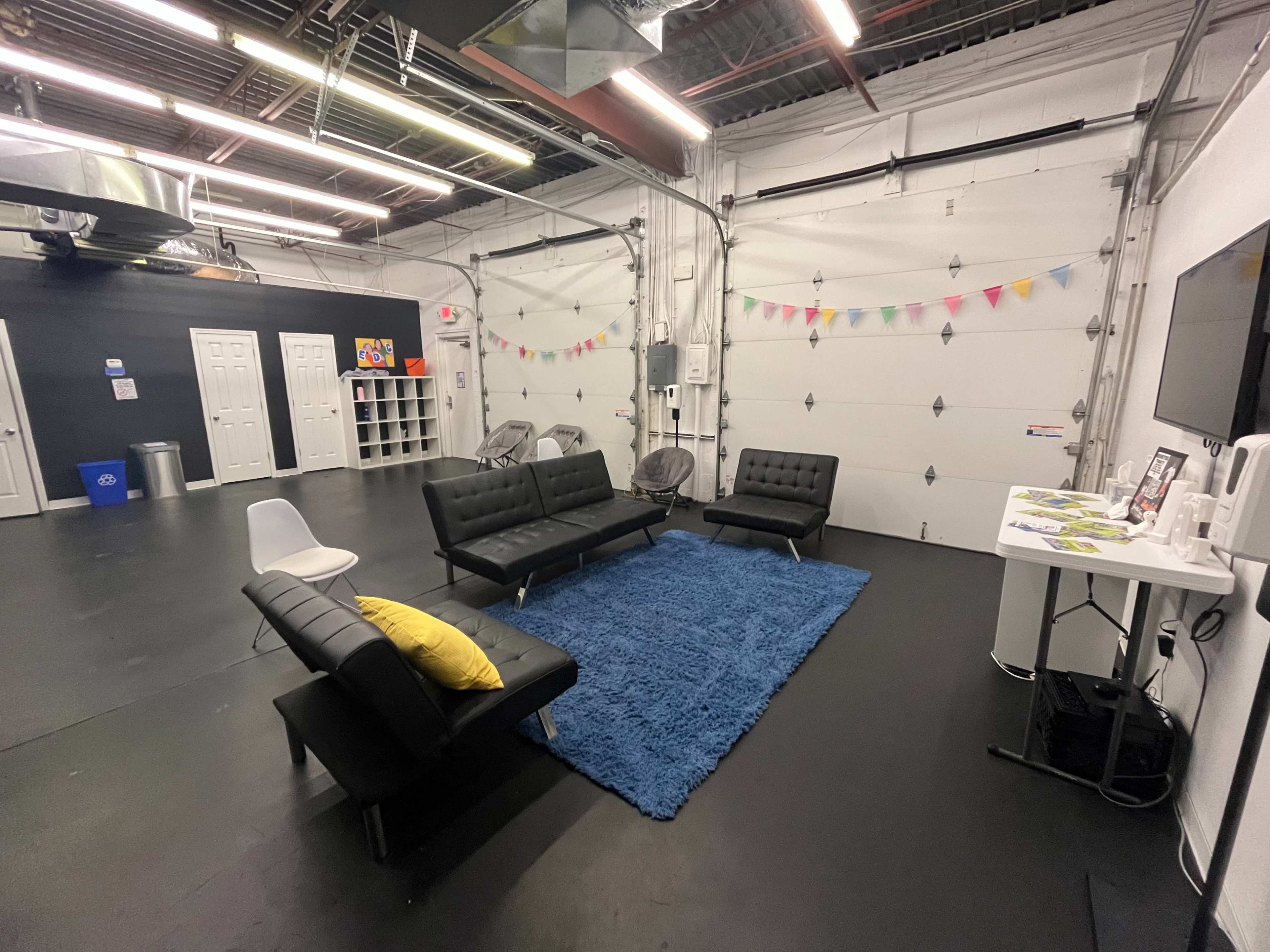 A modern lounge area features black couches, a blue rug, a white chair, and a wall-mounted TV in front of a gray wall with storage units and colorful bunting.