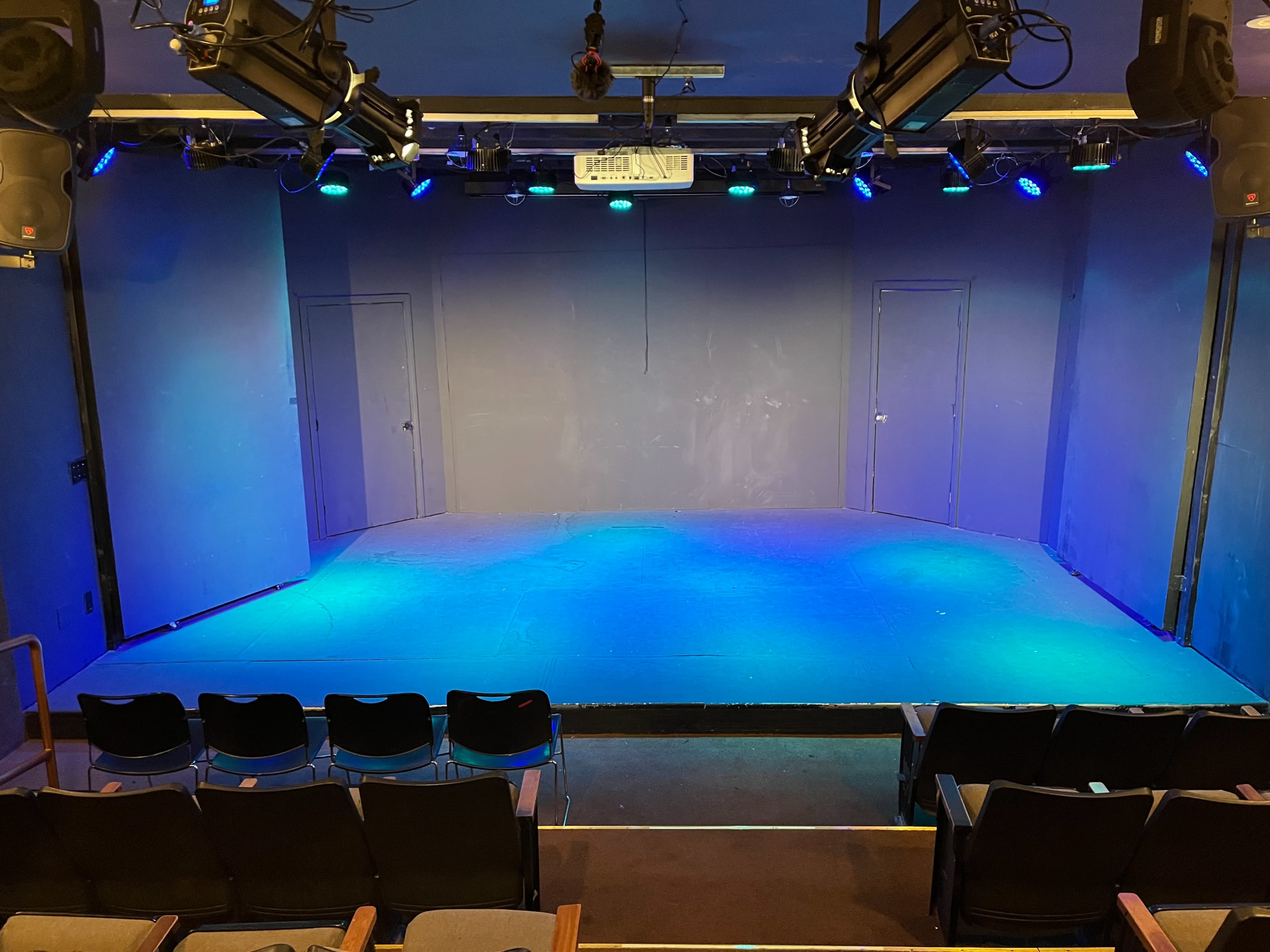 The image shows a small performance stage with blue lighting and empty seating arranged in front.