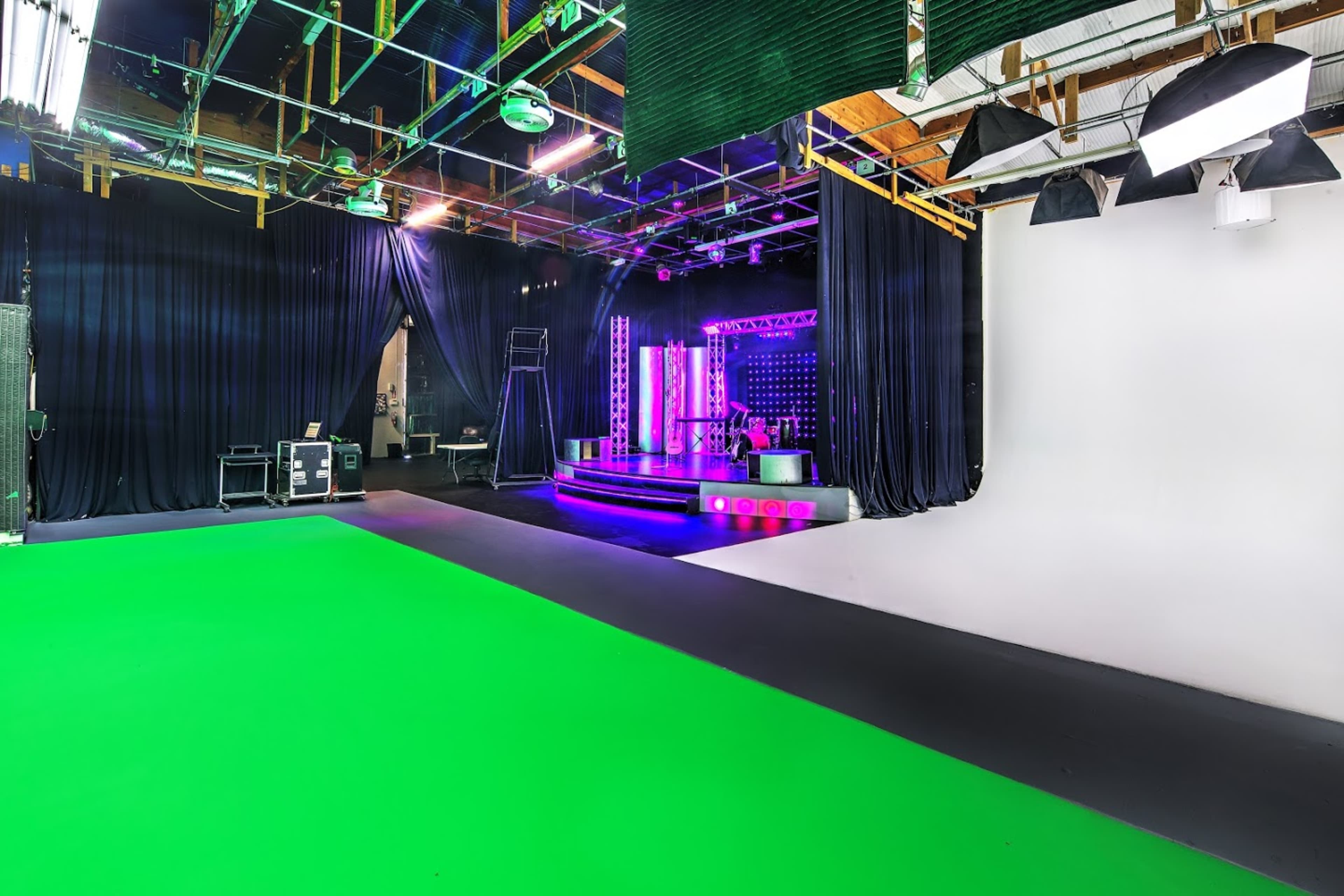 A production studio with a green screen area, black drapes, and a stage featuring colorful lights and equipment in the background.