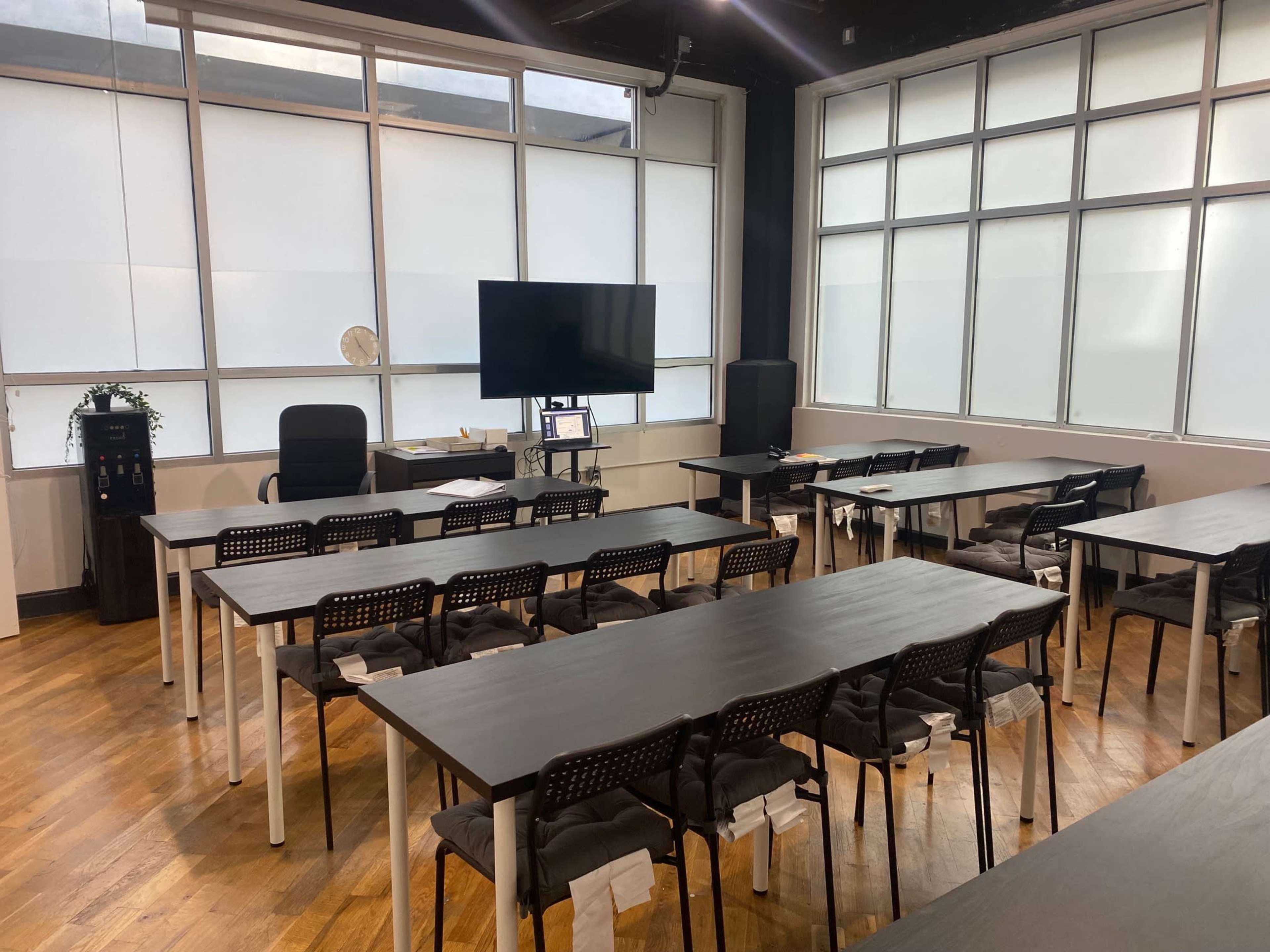 A well-lit classroom or meeting space with several tables and chairs arranged in a dual-row layout, a large screen on the wall, and a desk in the background.