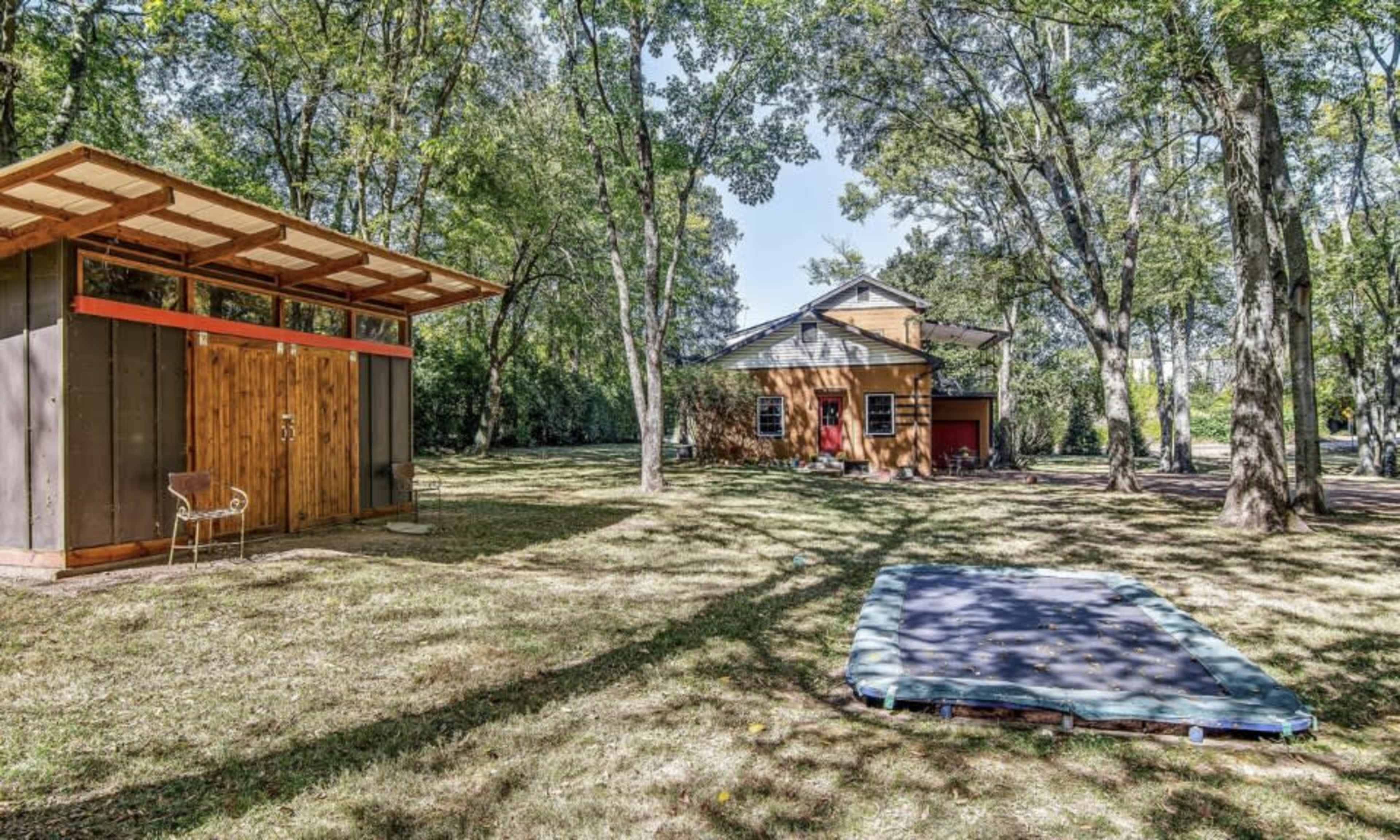 A wooden shed and a sleeping pad sit under trees in a grassy yard next to a house.