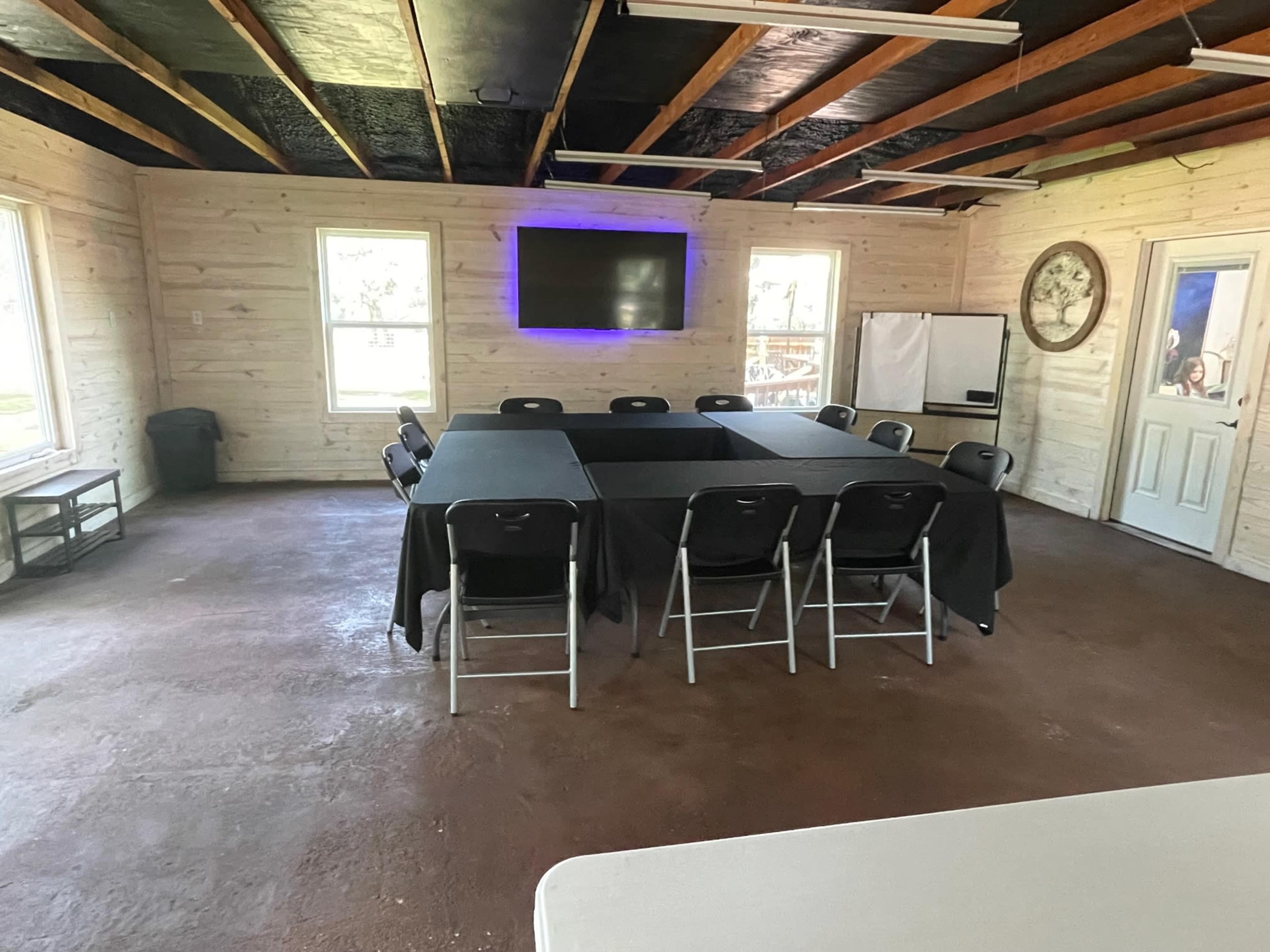 A meeting room features a square table surrounded by chairs, with a television mounted on the wall and windows allowing natural light.