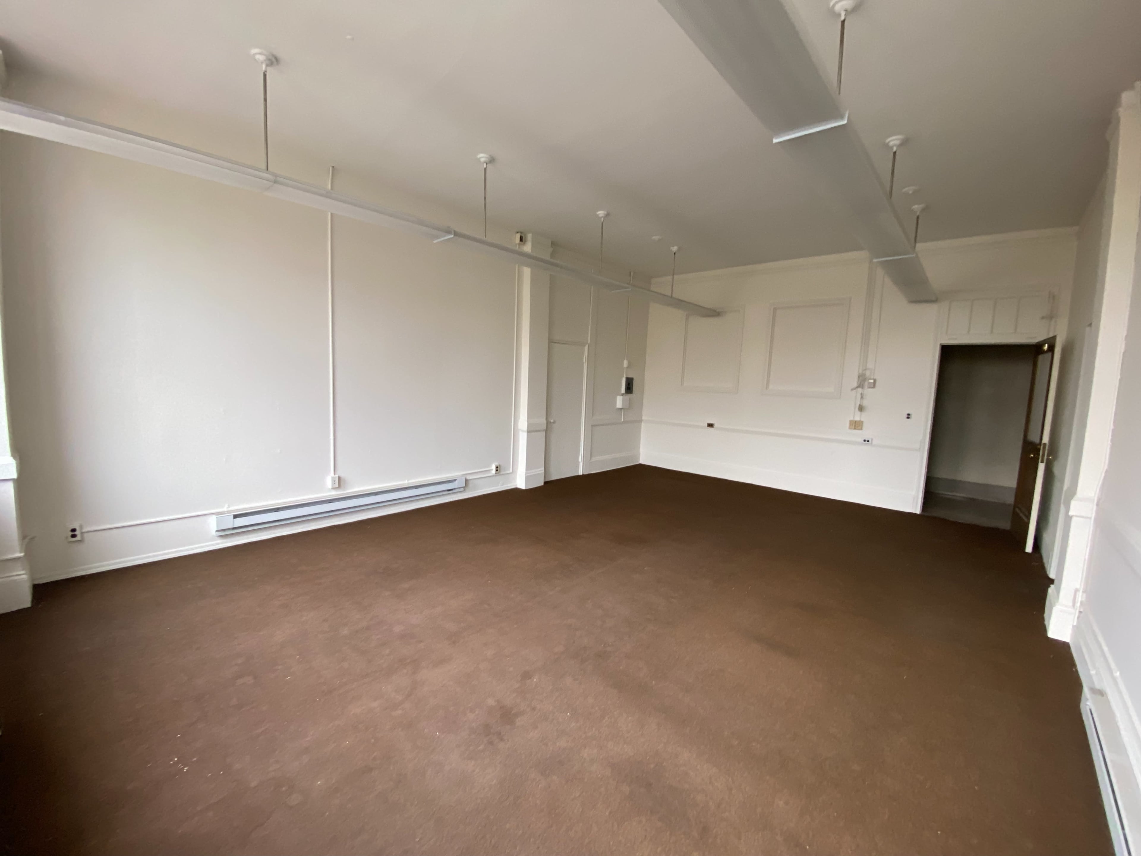 The image shows an empty room with brown carpeting, white walls, and overhead lighting fixtures.
