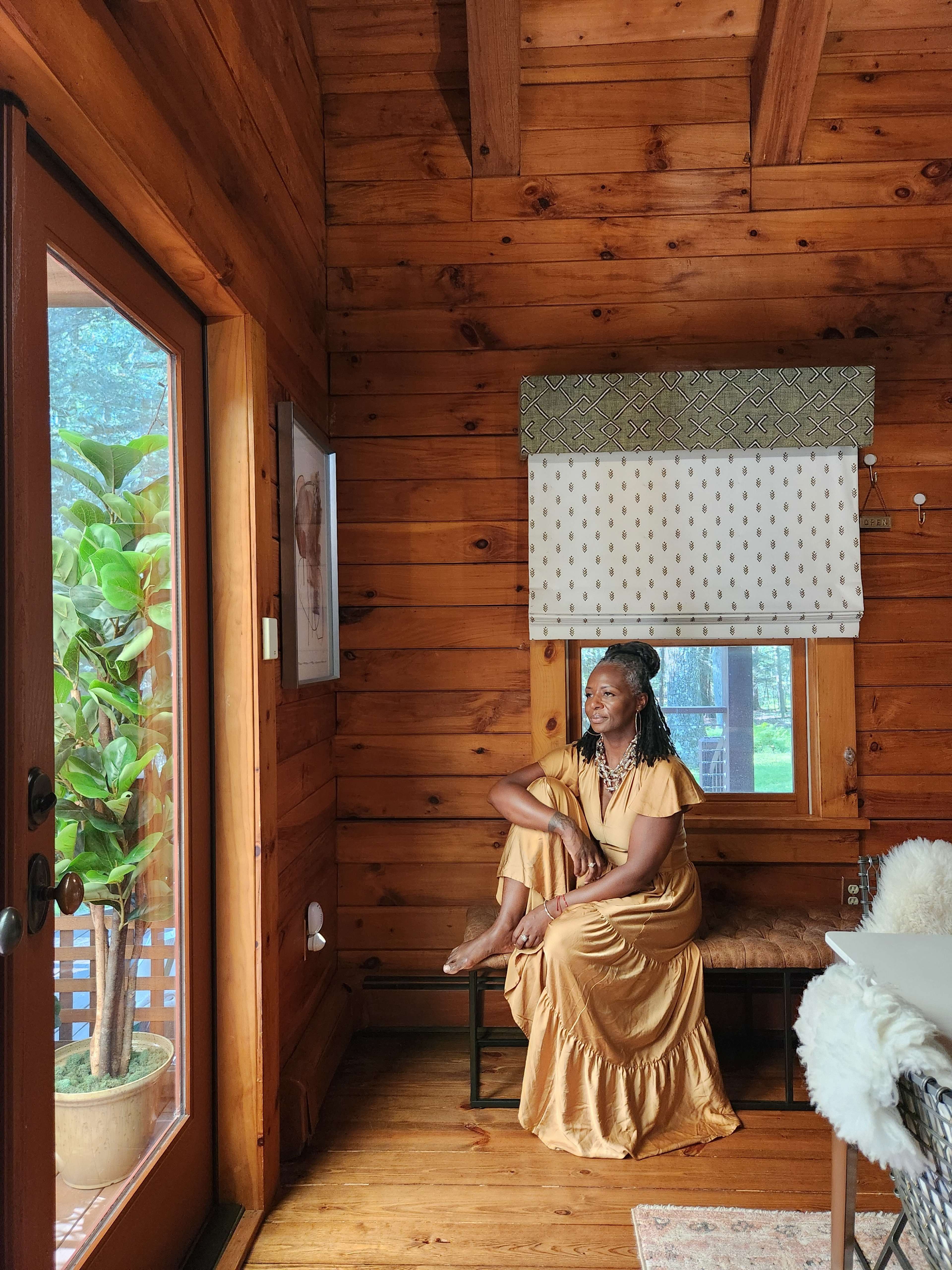 A woman with braids sits on a bench by a large window in a wooden cabin, with a potted plant nearby.