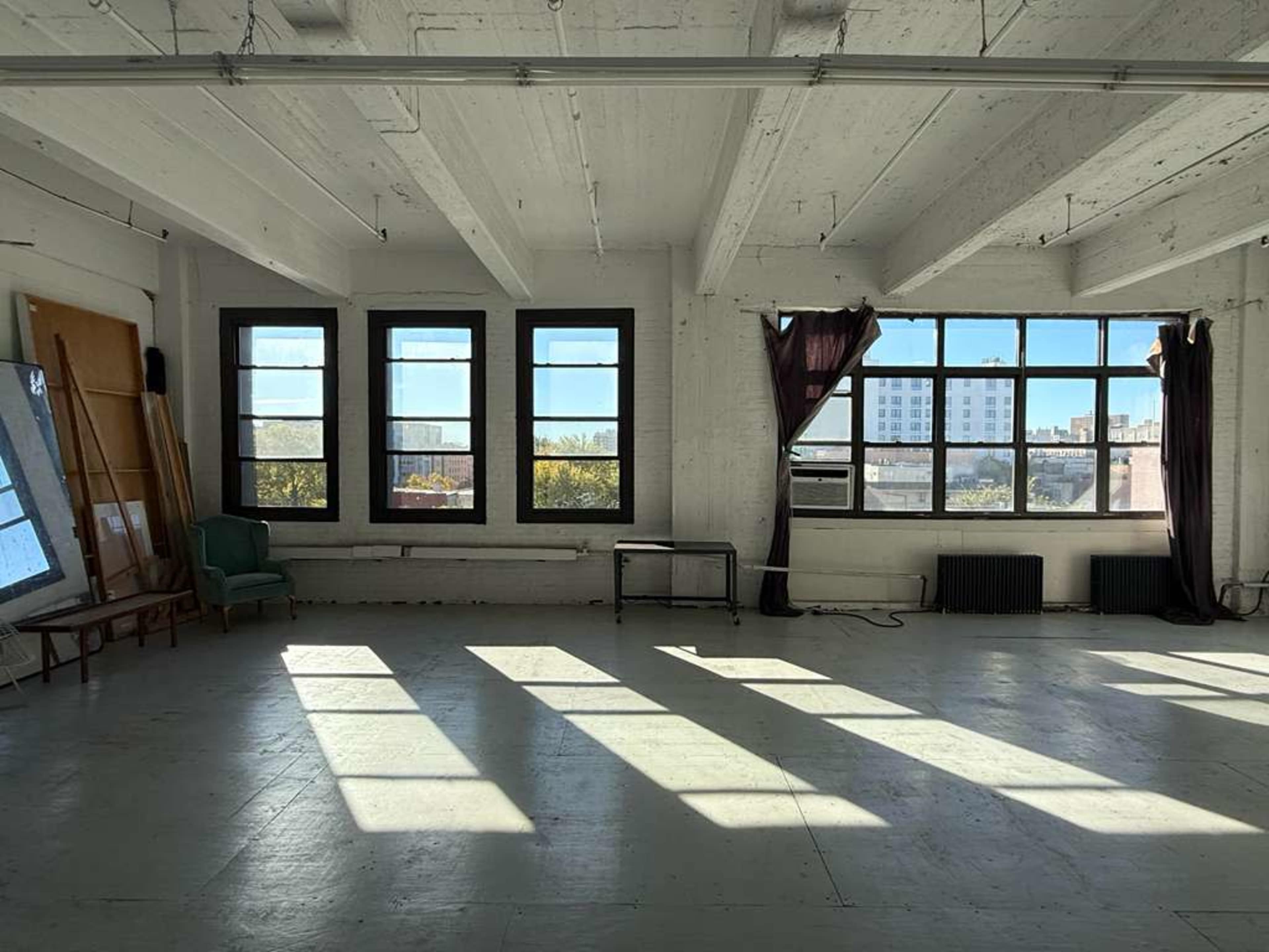 The image shows a spacious, well-lit room with large windows casting shadows on the white floor, featuring a green chair and a folding table against the wall.