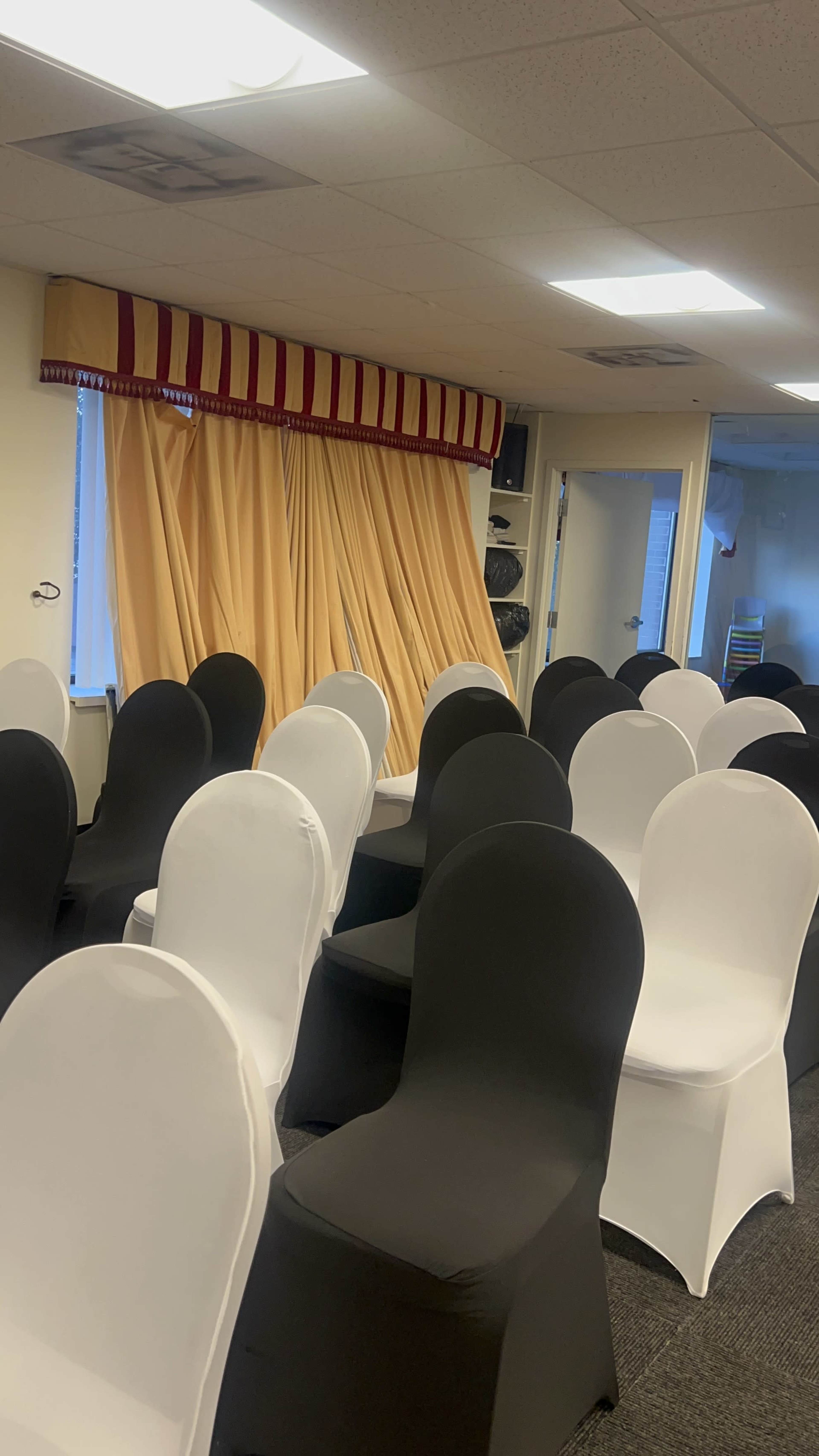 A room is set up with rows of black and white chairs facing a window with yellow curtains.