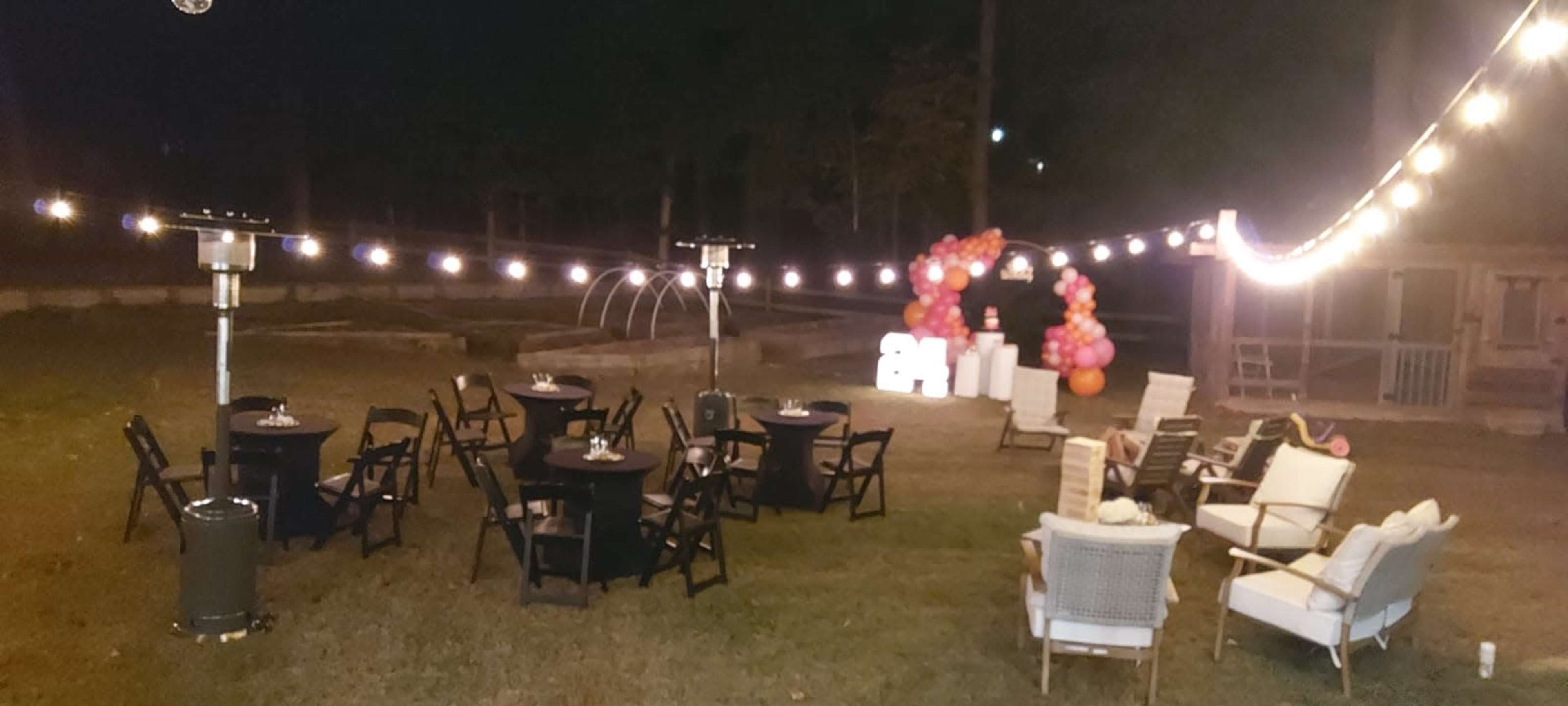 An outdoor party setup at night with seating arrangements, decorative lights, and a balloon arch in the background.