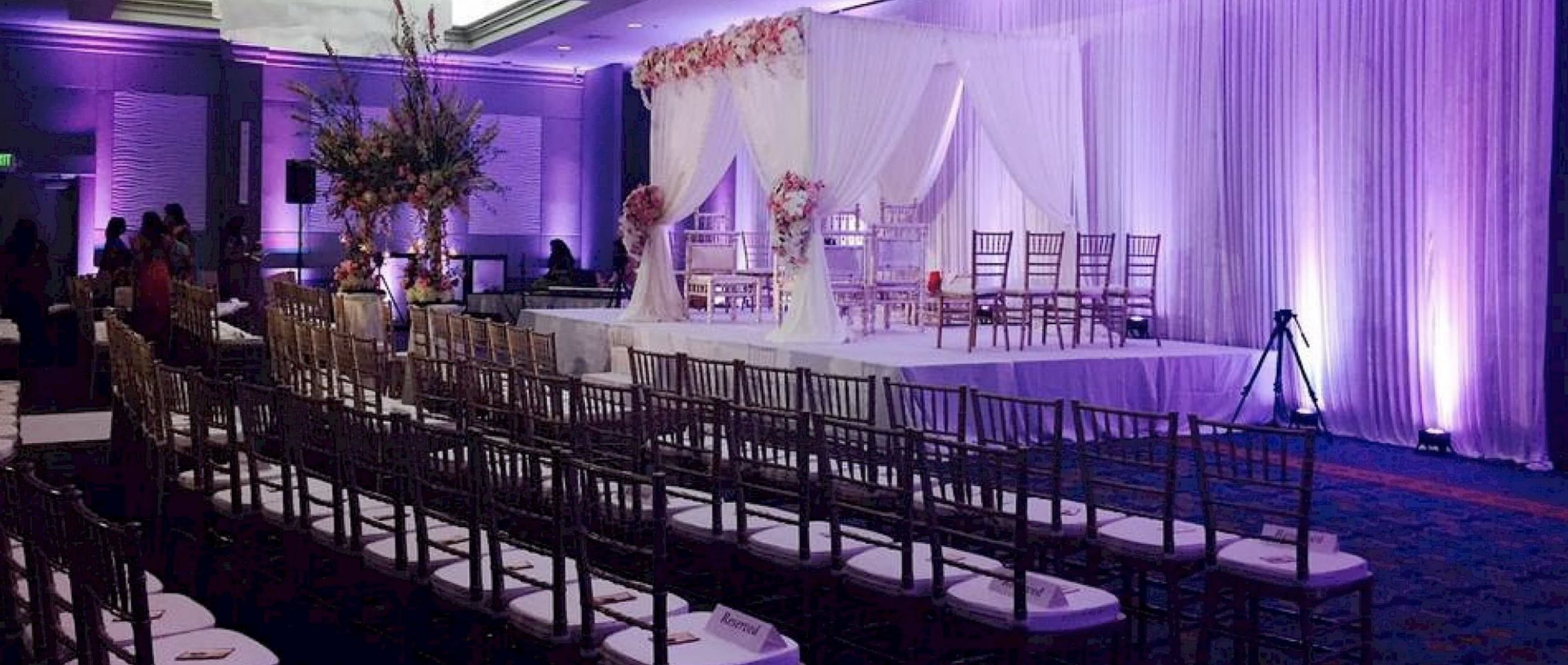 The image shows a decorated event space featuring a stage with floral arrangements, white drapery, and rows of chairs arranged in front of it.