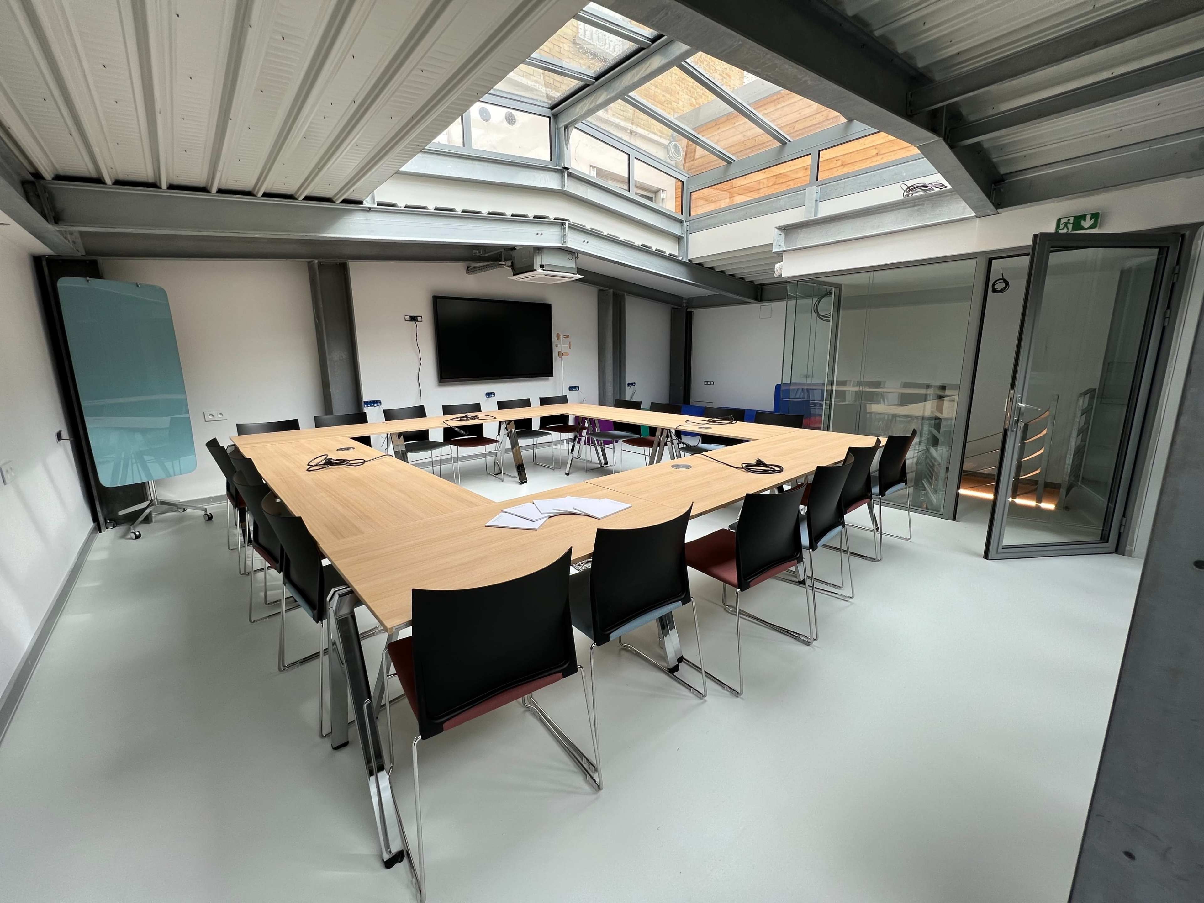 A modern conference room features a large rectangular table surrounded by black chairs, with a screen mounted on the wall and large windows above.