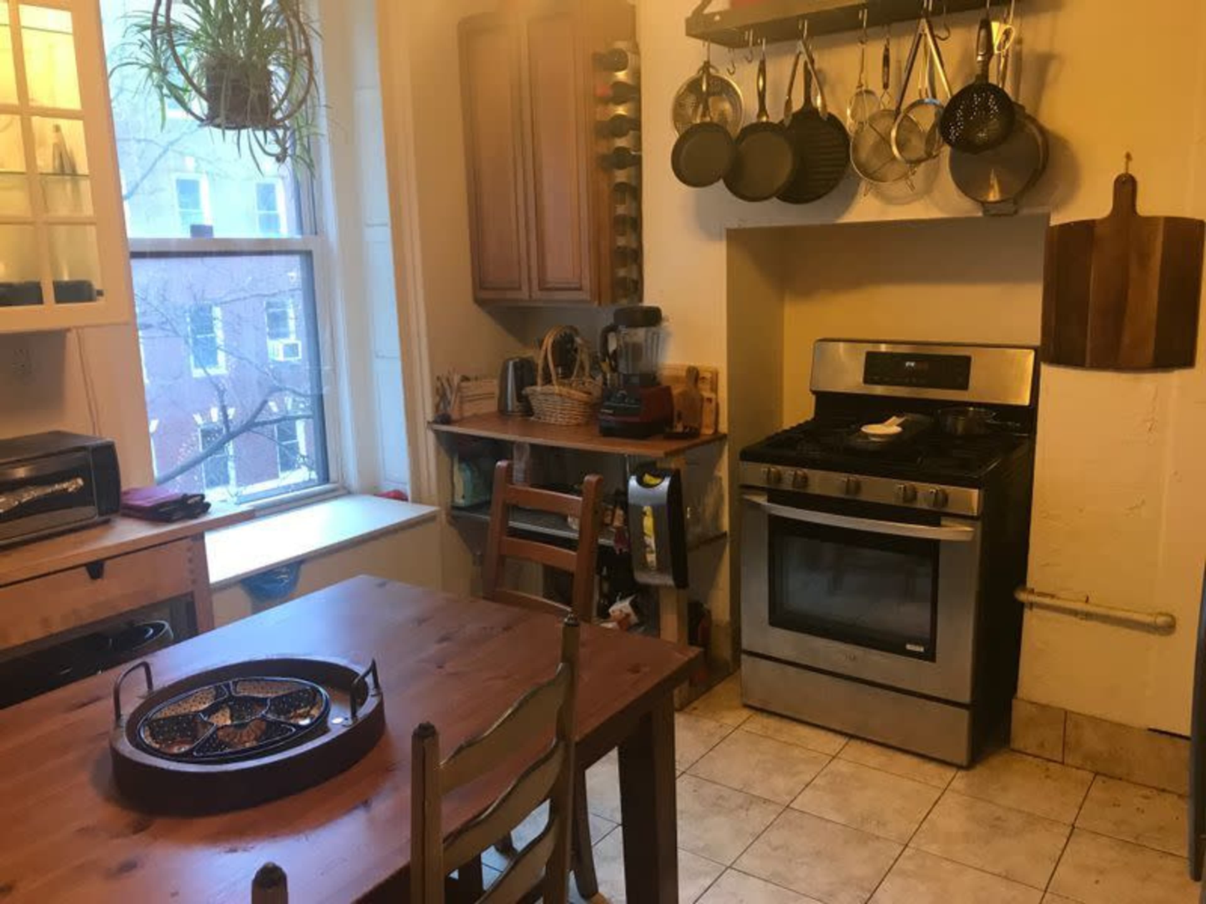 The image shows a kitchen with a wooden table, an arranged countertop with cookware, and a window allowing natural light.