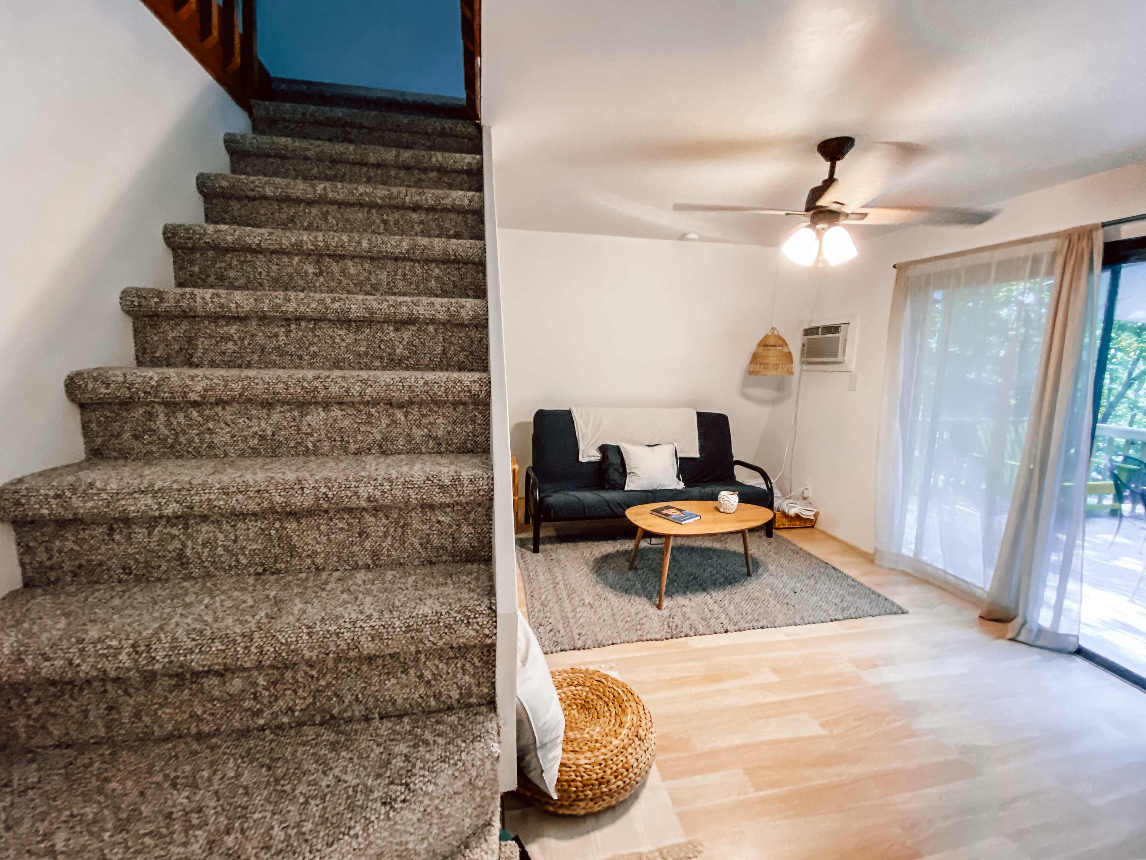 The image shows a living area with a sofa and coffee table, adjacent to a carpeted staircase leading to a second floor.