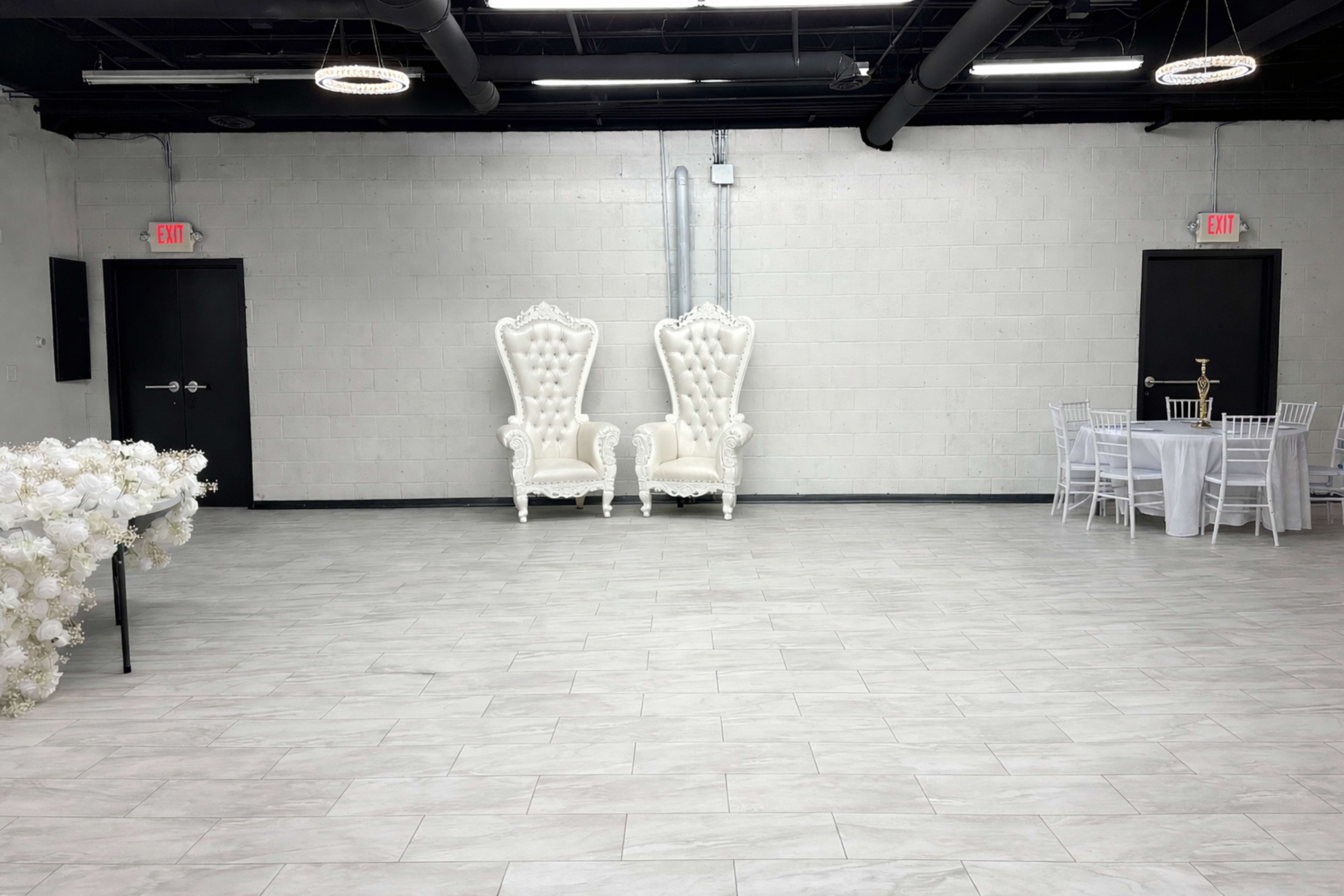 The image shows a spacious event venue with white tiled floors, two ornate white armchairs, a round table set for dining, and floral arrangements on one side.