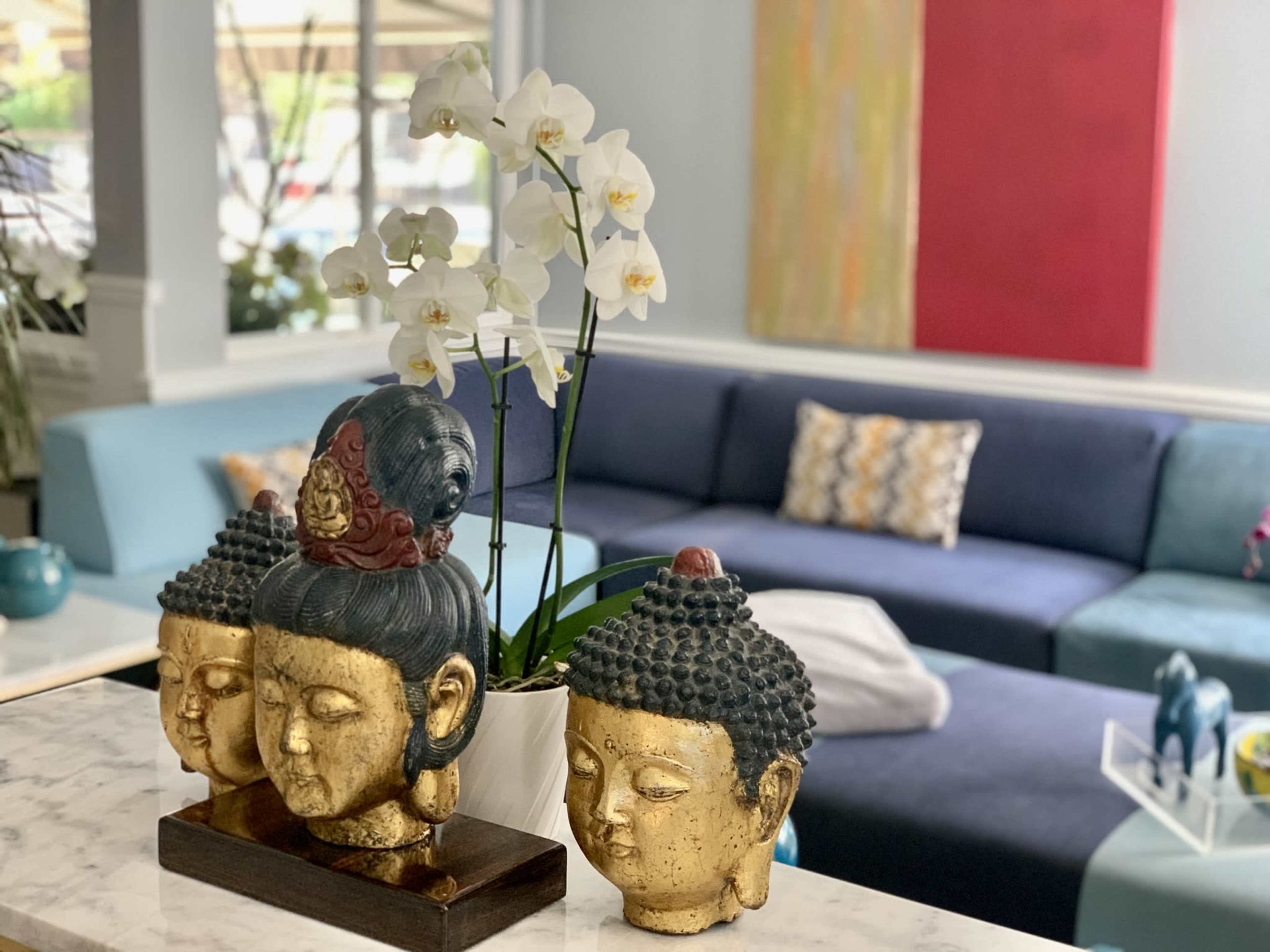 A set of decorative Buddha sculptures and a white orchid plant are placed on a marble surface in a room featuring a blue sofa and colorful wall art.