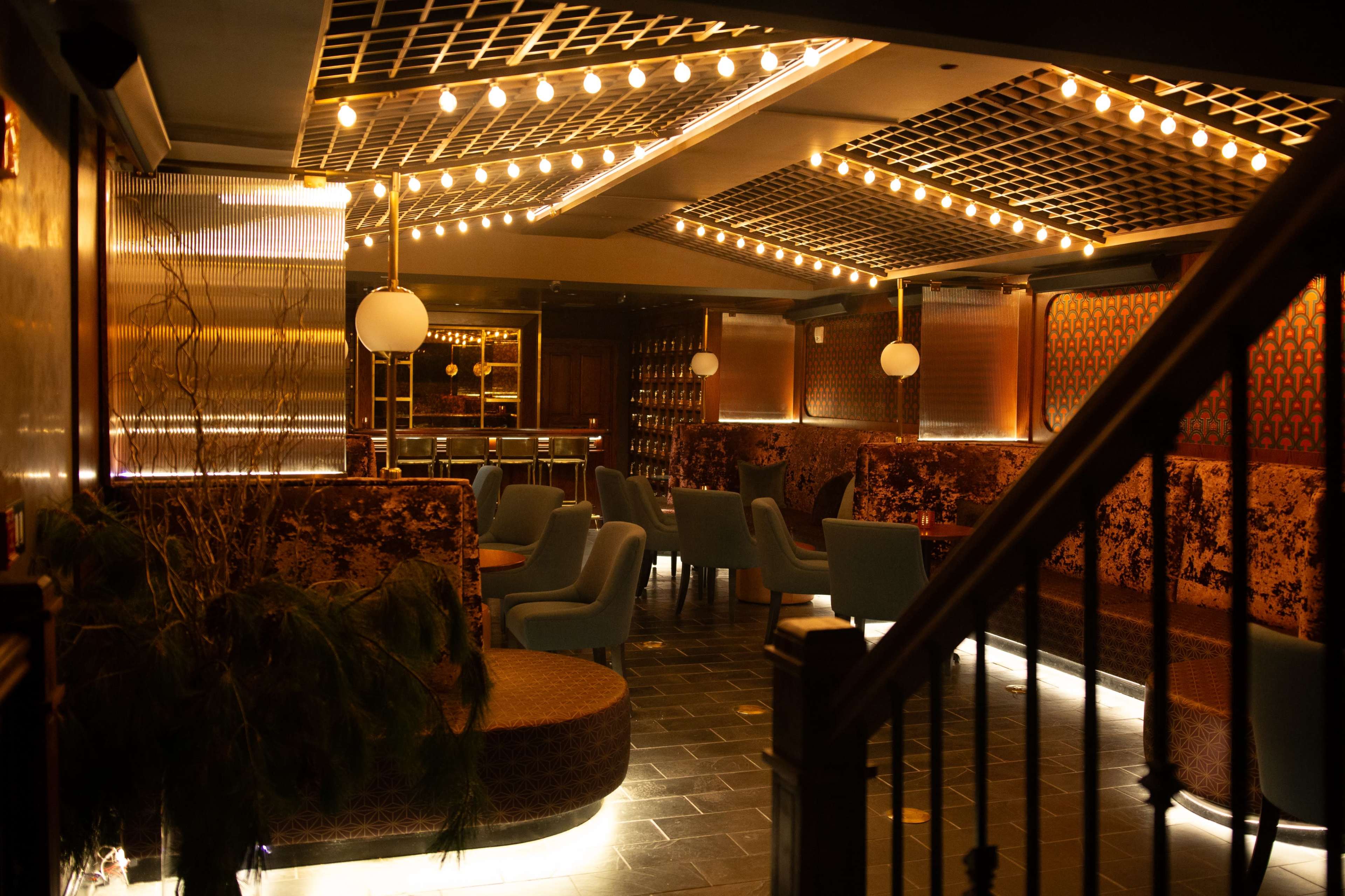 The image shows a dimly lit lounge with plush seating, decorative lighting, and a stylish bar area in the background.