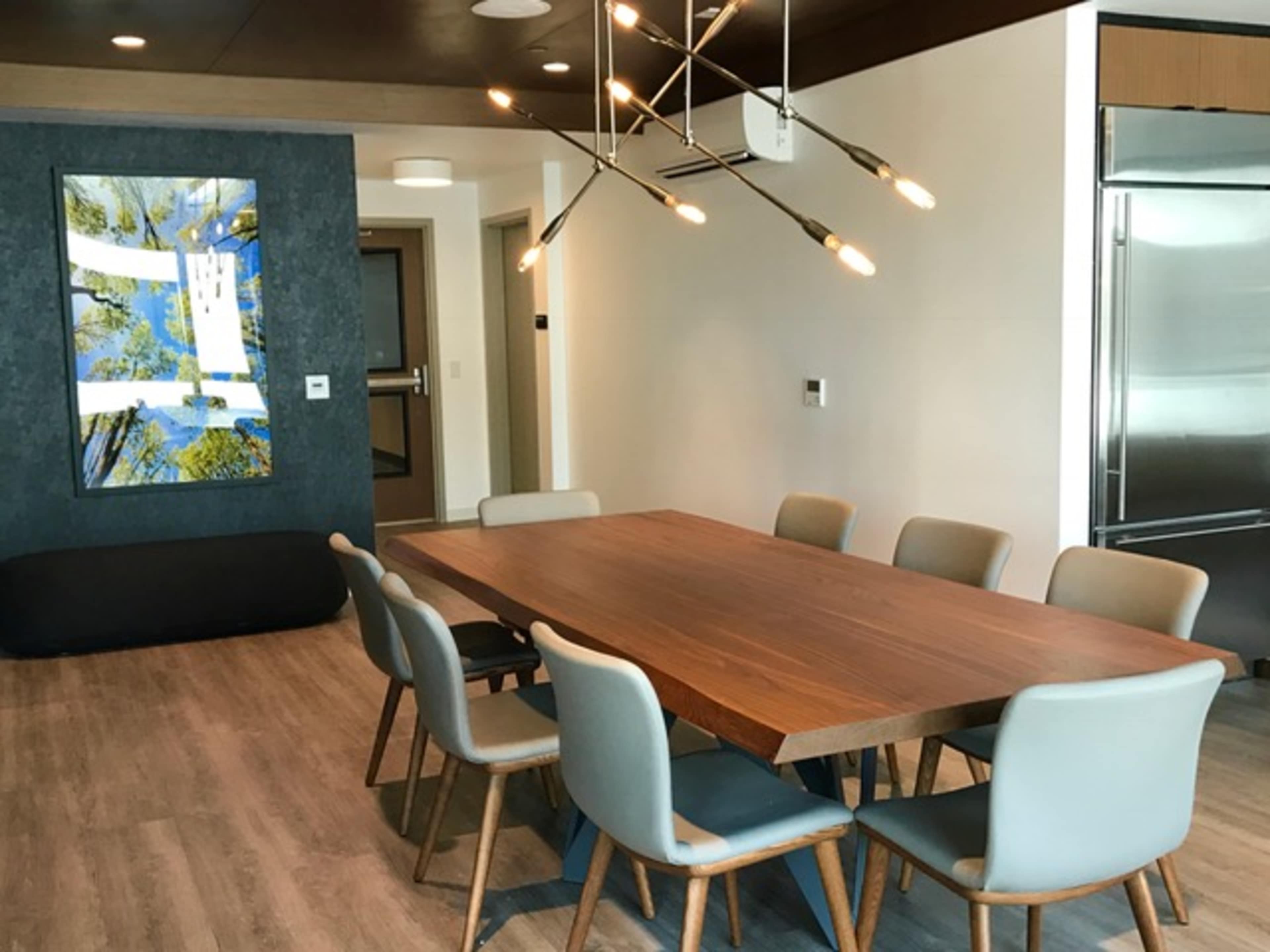 A modern dining area features a large wooden table surrounded by eight chairs, with a decorative light fixture overhead and an artwork displayed on the wall.