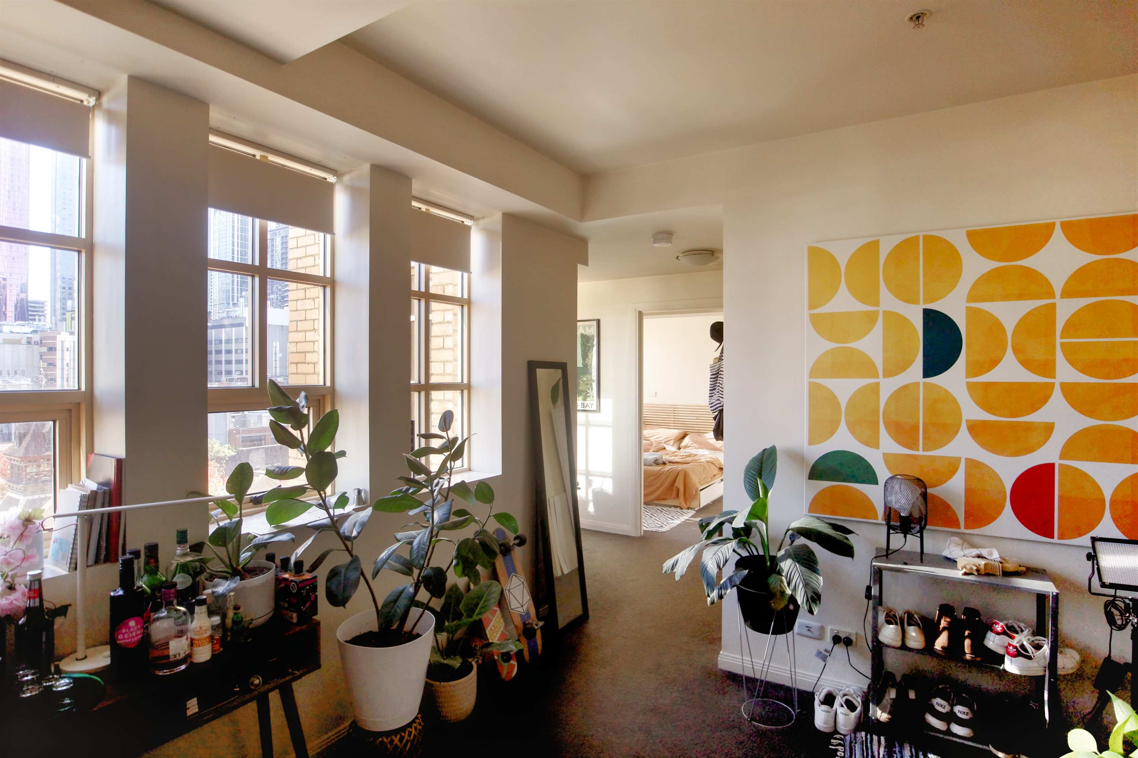 A bright interior of a room featuring large windows, plants, a mirror, a colorful geometric wall art, and a glimpse of a bedroom in the background.