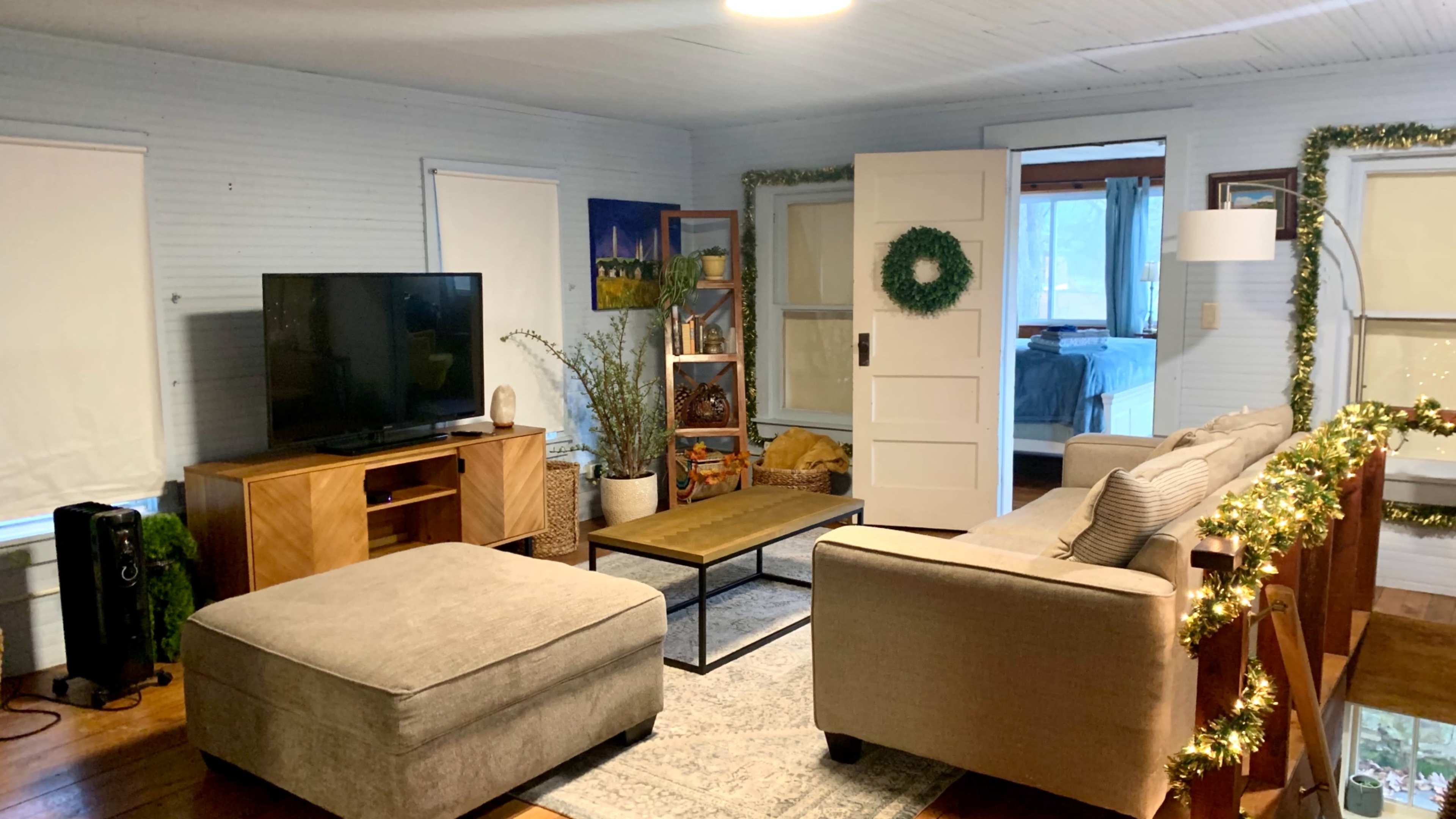 The image shows a living room with a television on a wooden console, a beige sofa and ottoman, a coffee table, and decorative greenery along the walls and furniture.