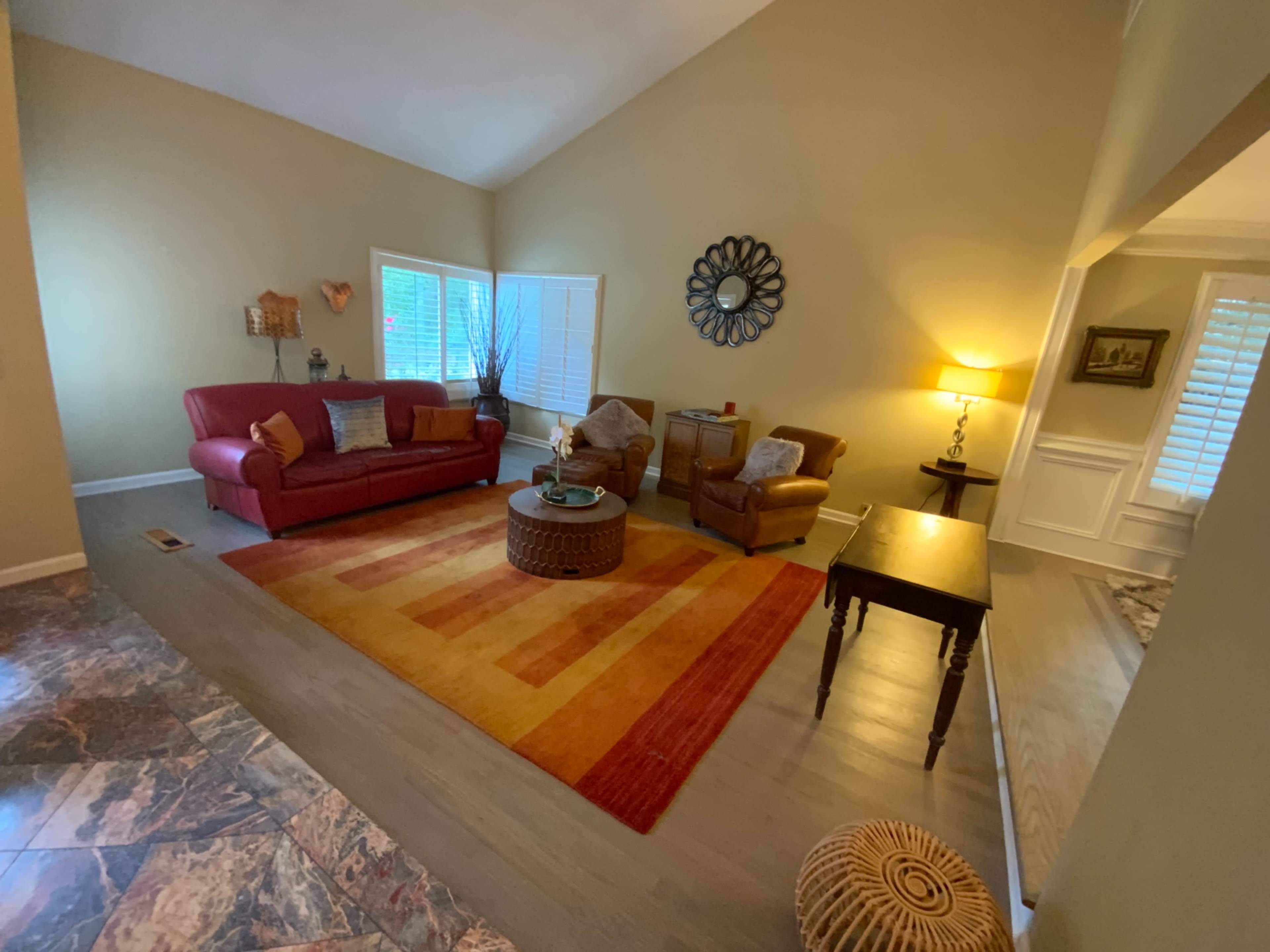A cozy living room with a red sofa, two brown armchairs, a round coffee table on a colorful rug, and a small side table, all illuminated by a lamp in the corner.