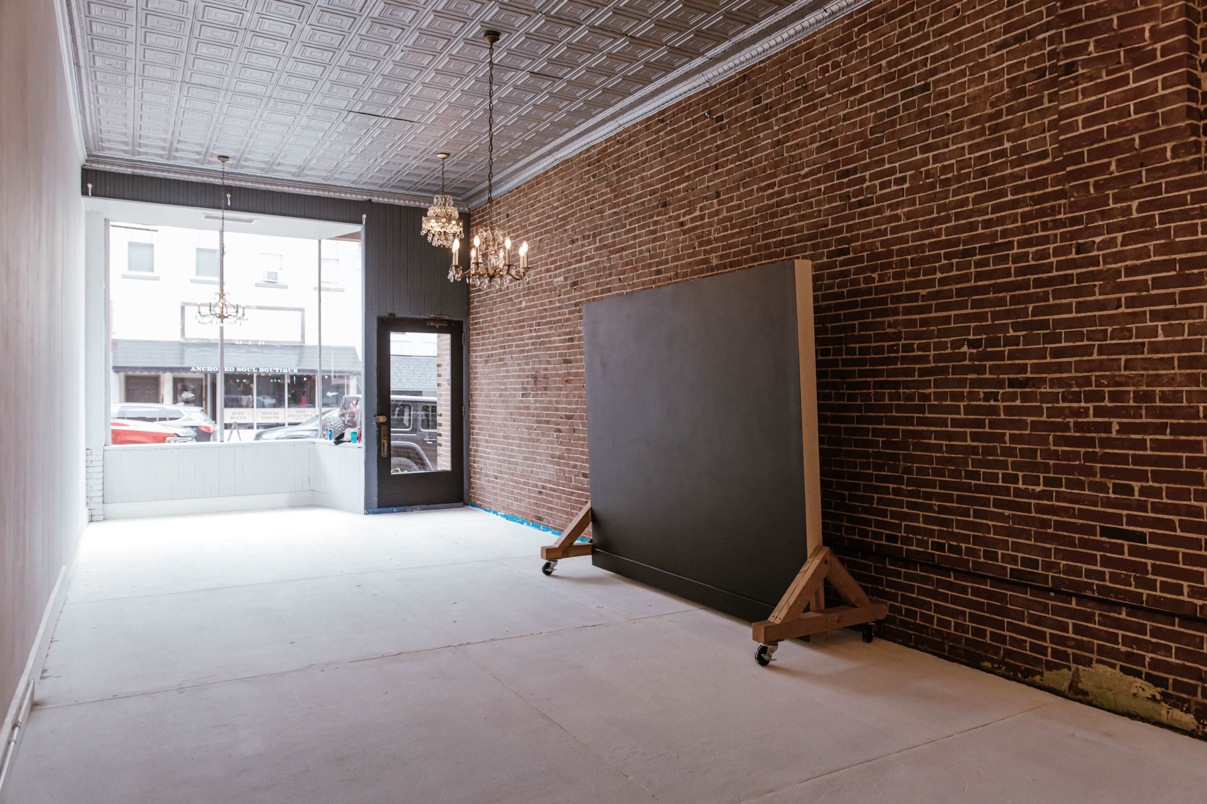 The image shows an empty room with exposed brick walls, a large movable panel on wheels, and a window facing a street.