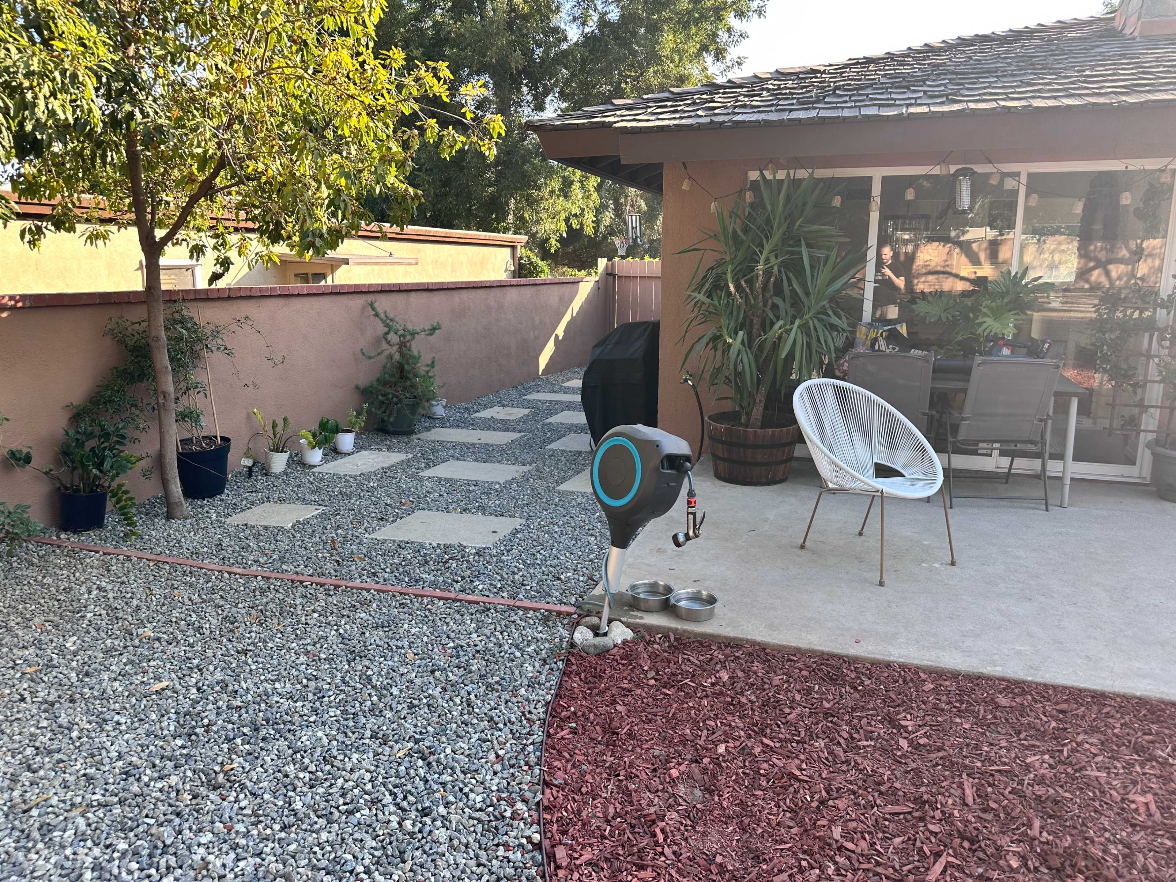 The image shows a landscaped backyard area featuring a stone pathway, plants along the edges, a seating arrangement, and a pet bowl station.