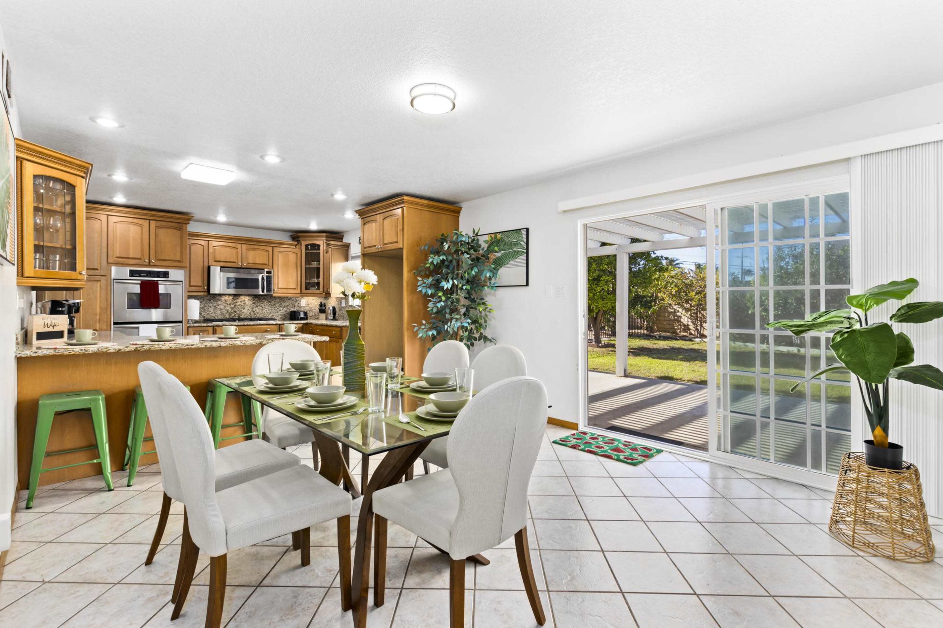 A spacious kitchen and dining area feature a glass-top table set for six, surrounded by light-colored chairs, with wooden cabinets and modern appliances in the background.